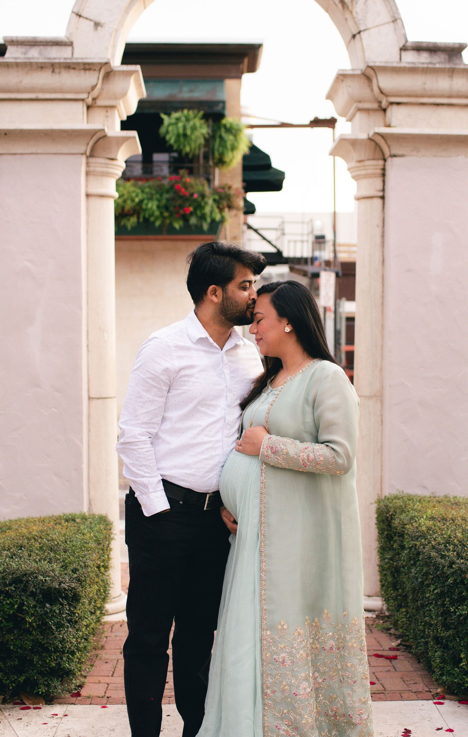 A couple in traditional attire share an intimate moment under decorative stone arches in a garden setting.
