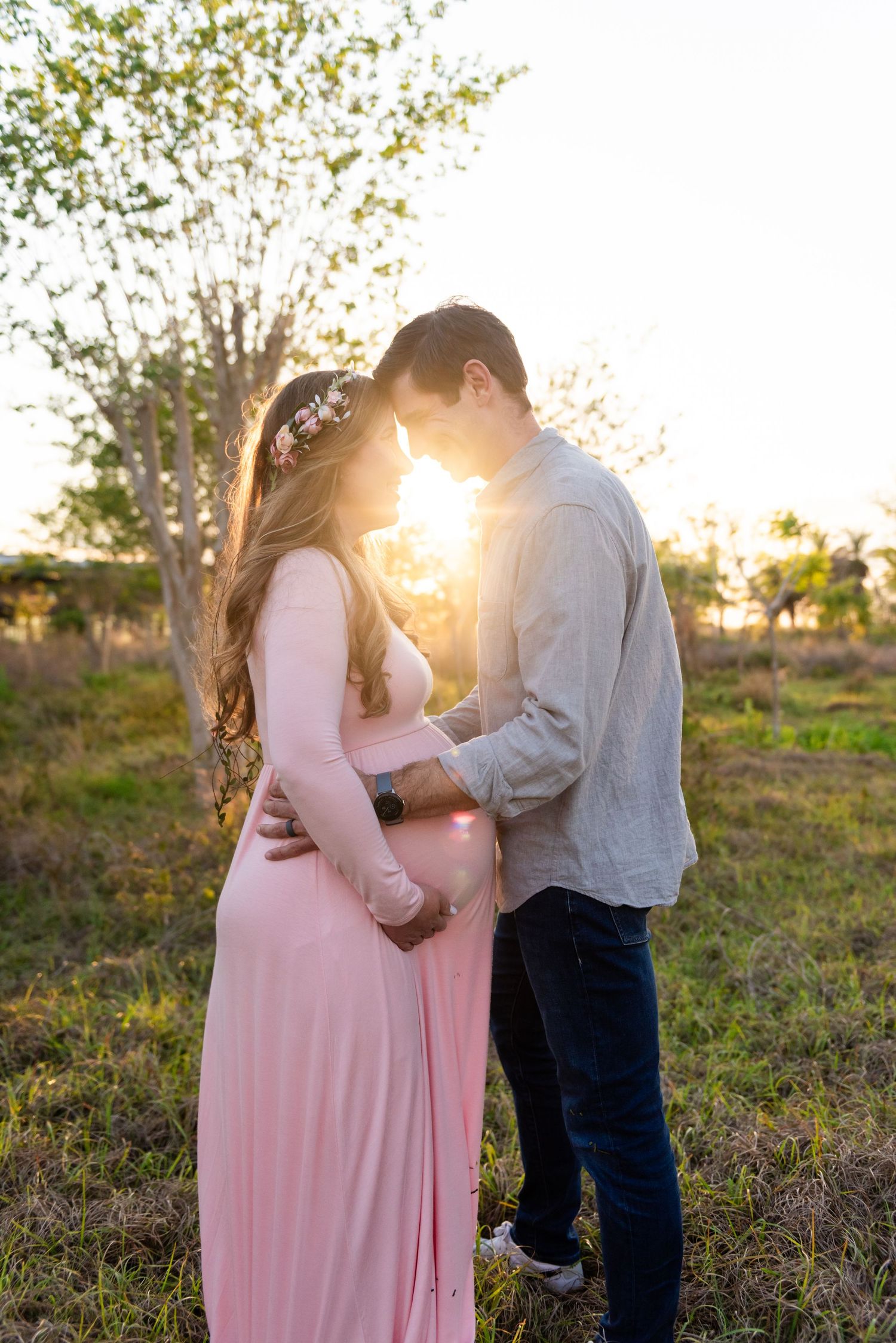 A couple in an outdoor setting at sunset, with the pregnant woman wearing a pink maxi dress while sharing an intimate moment.