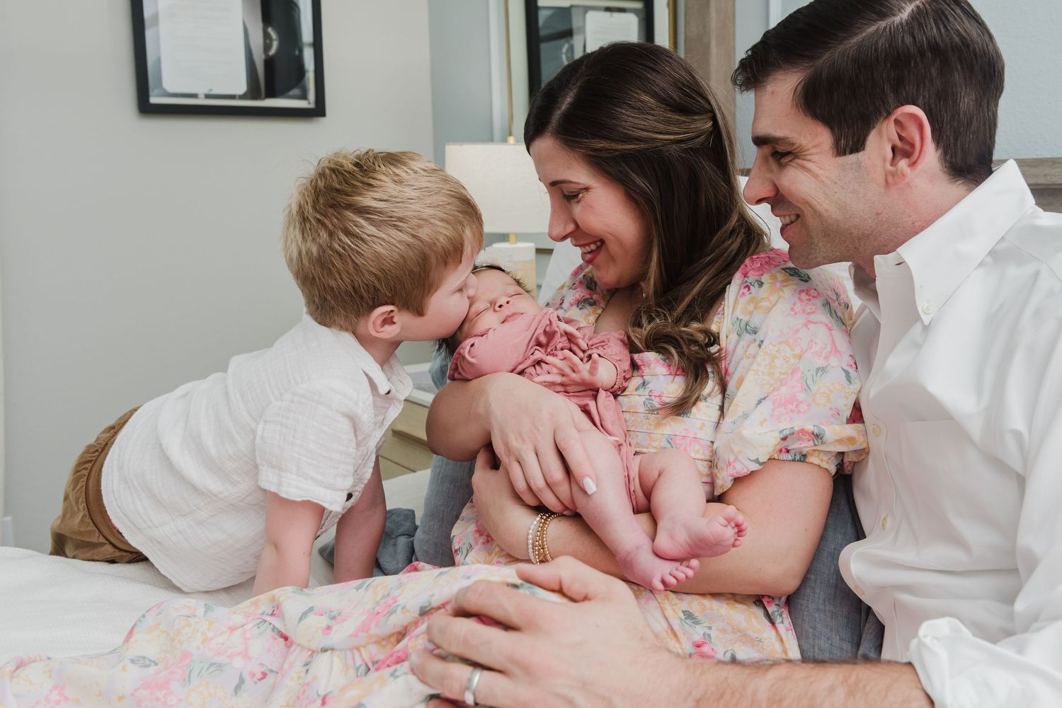 Intimate family moment as toddler leans in to kiss newborn sibling while parents watch lovingly.