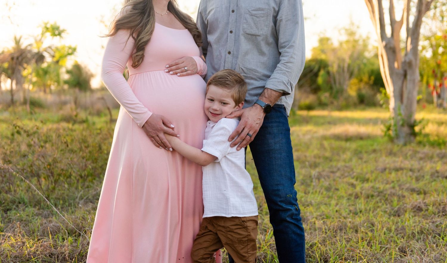 A pregnant woman in a pink dress stands outdoors with a young child at sunset in a natural setting.