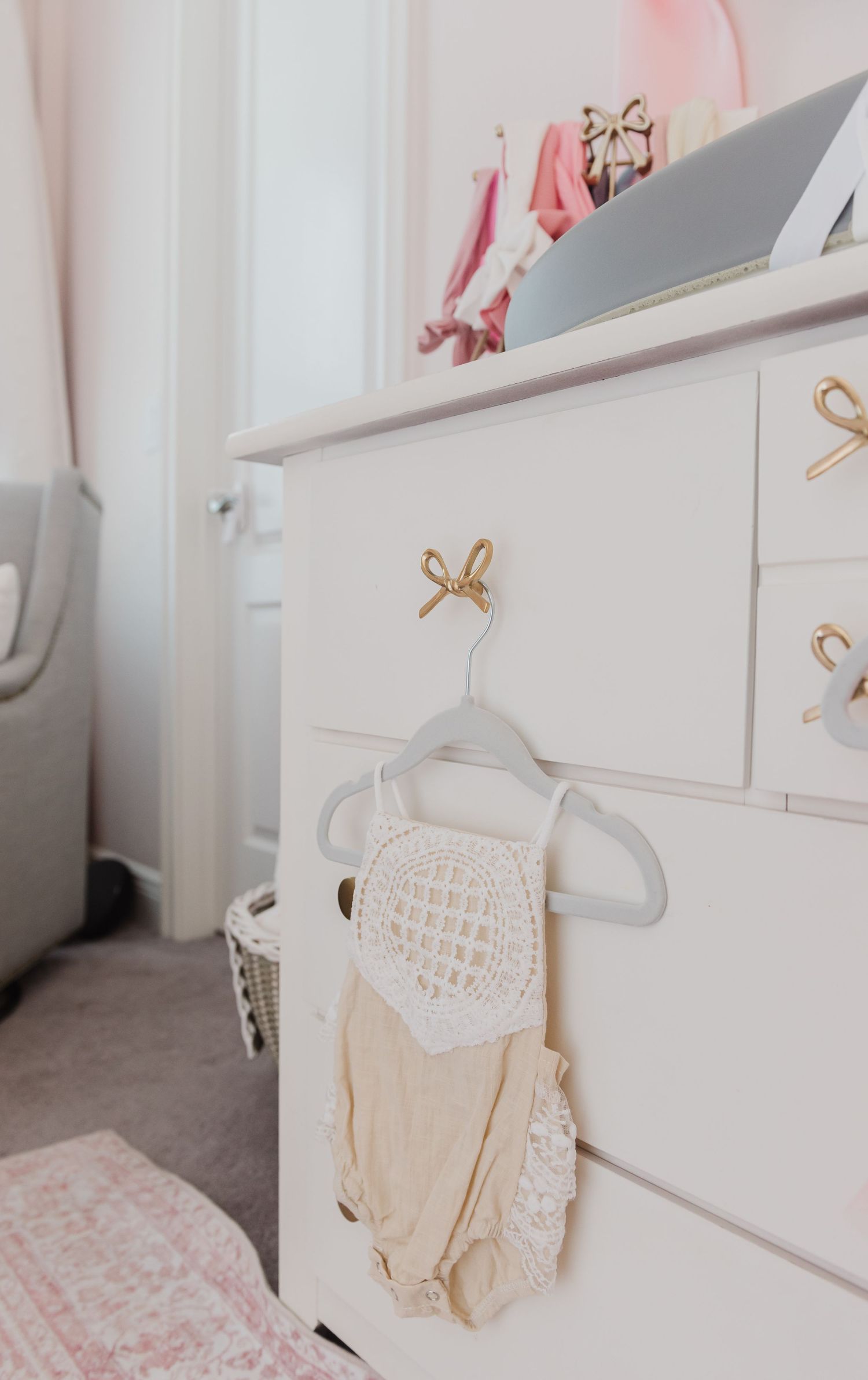White dresser with gold bow handles and baby clothing hanging from drawer in a nursery room.