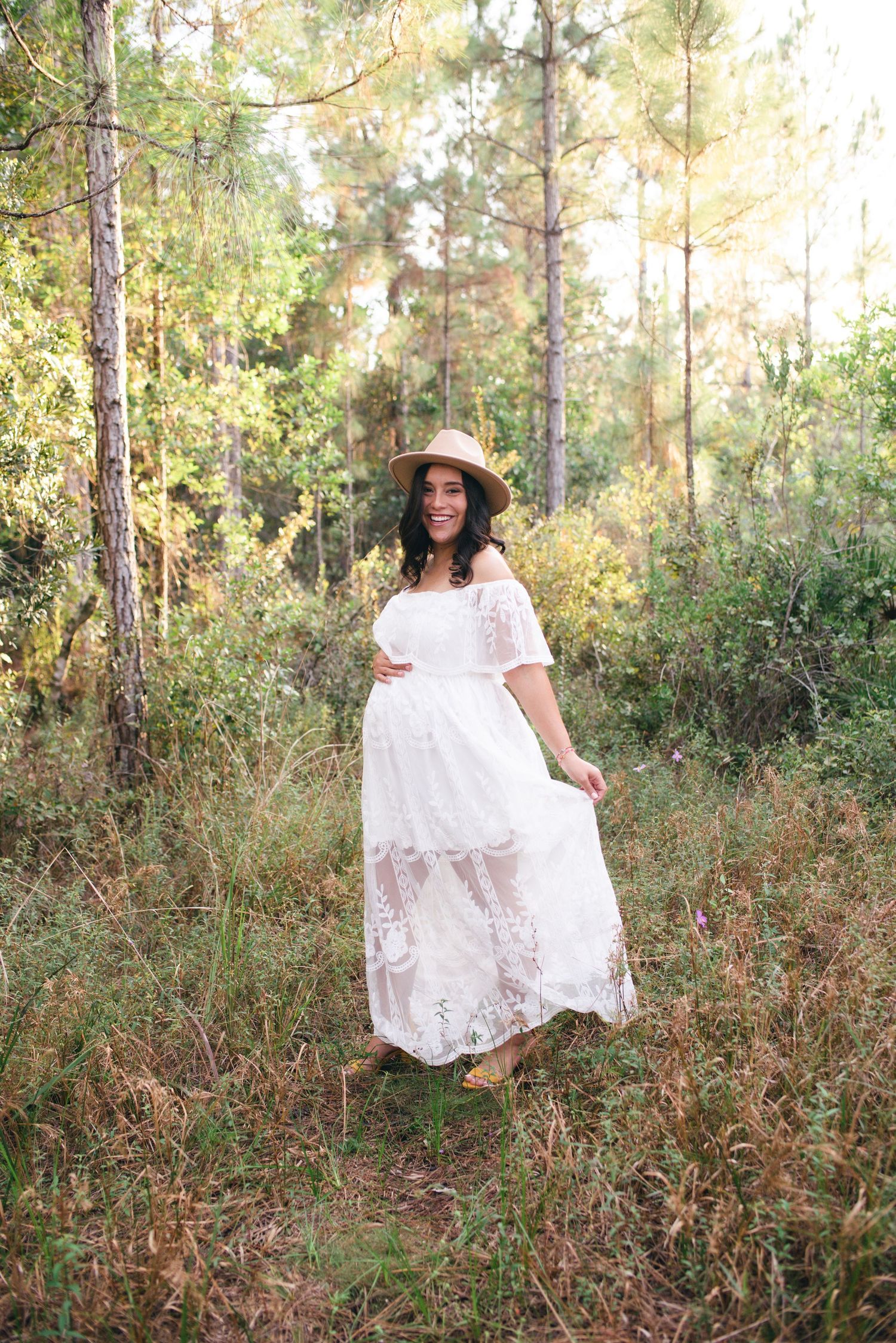 A person in a flowing white lace maternity dress and straw hat poses elegantly among tall pine trees in a golden forest.