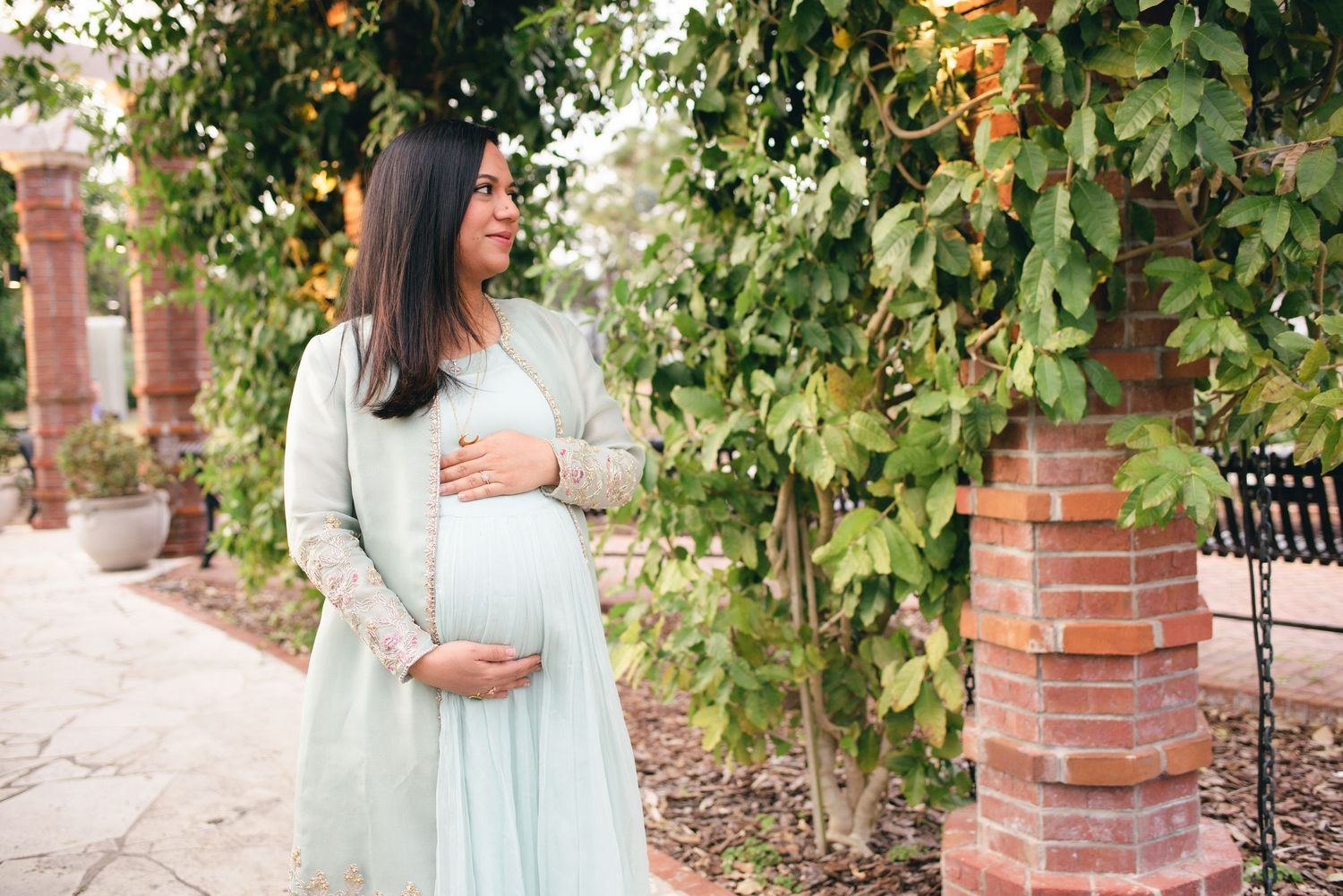 Expectant woman in a flowing mint green dress stands in a garden walkway with brick pillars and lush greenery.