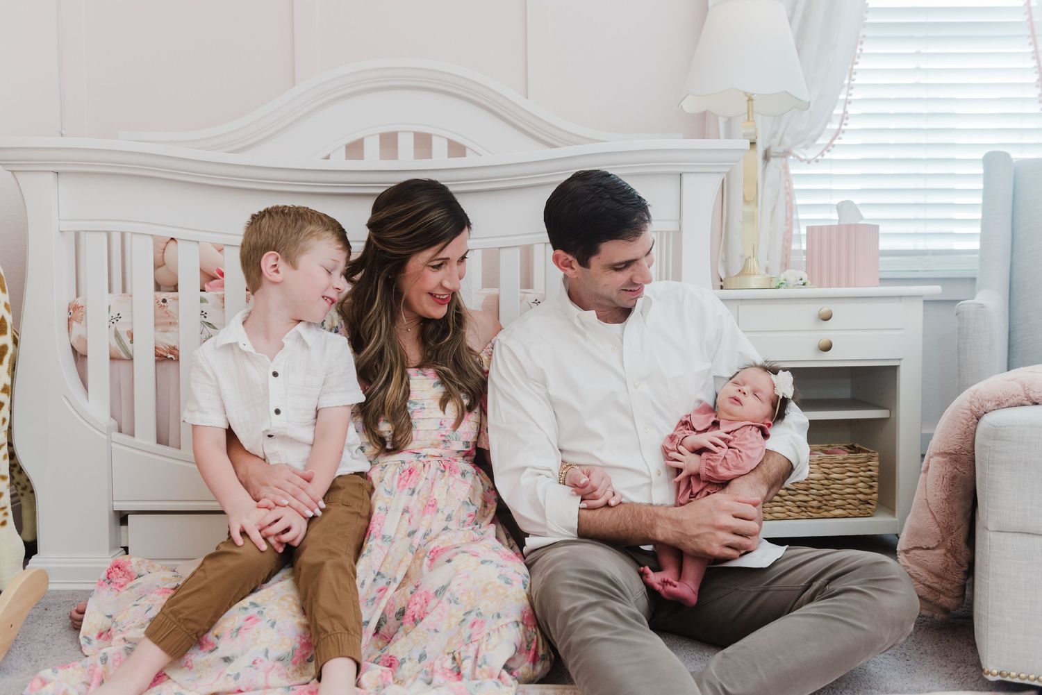 Family in coordinating white and pink outfits sits together on floor with newborn baby in nursery setting.
