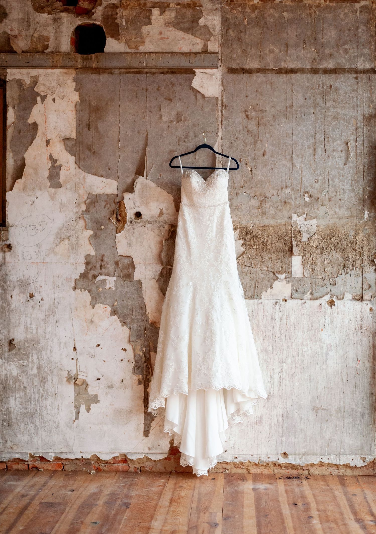 Elegant white wedding dress hanging against distressed wall in rustic room.