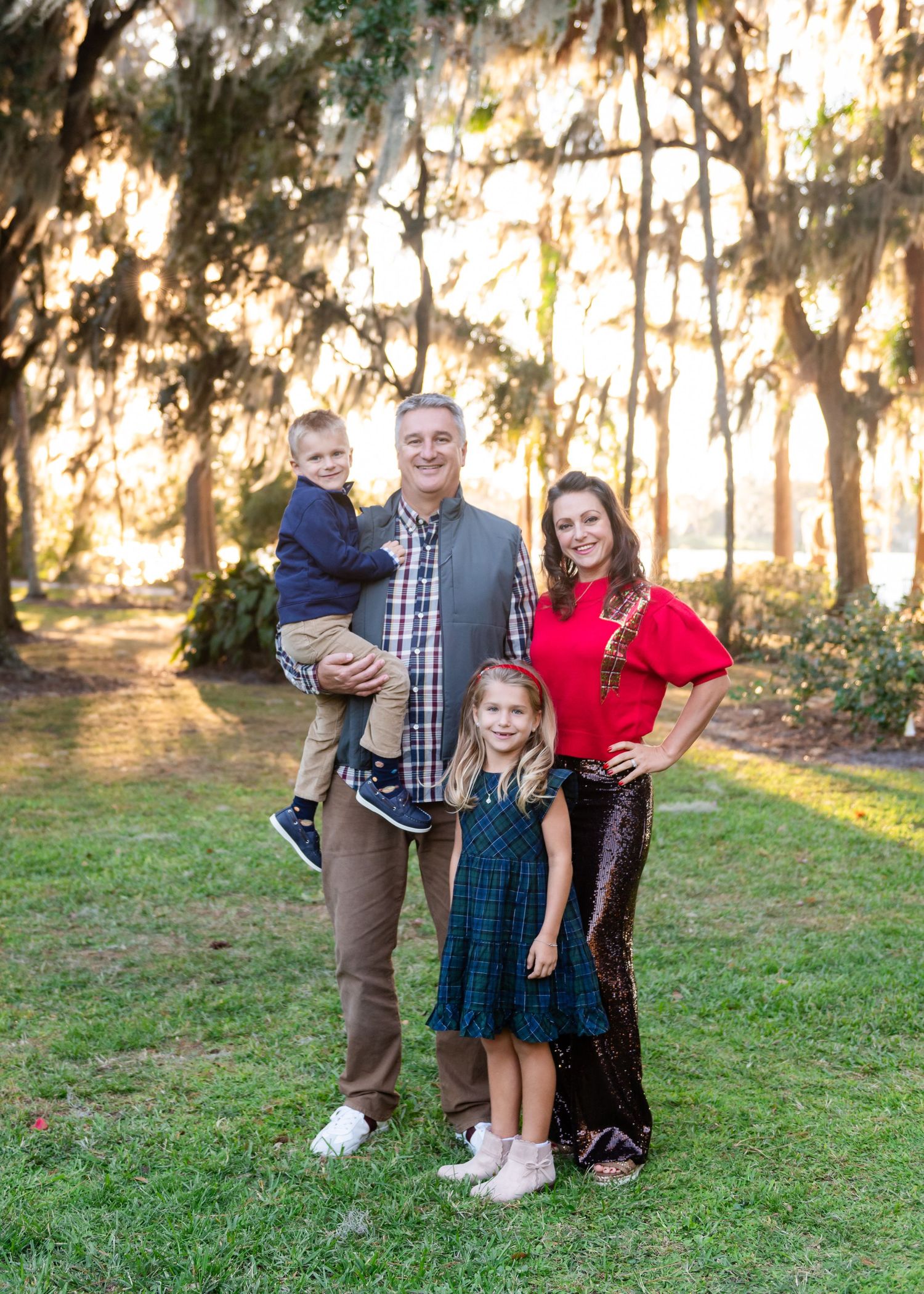 Family poses together outdoors at sunset with sunlit trees and Spanish moss in the background.