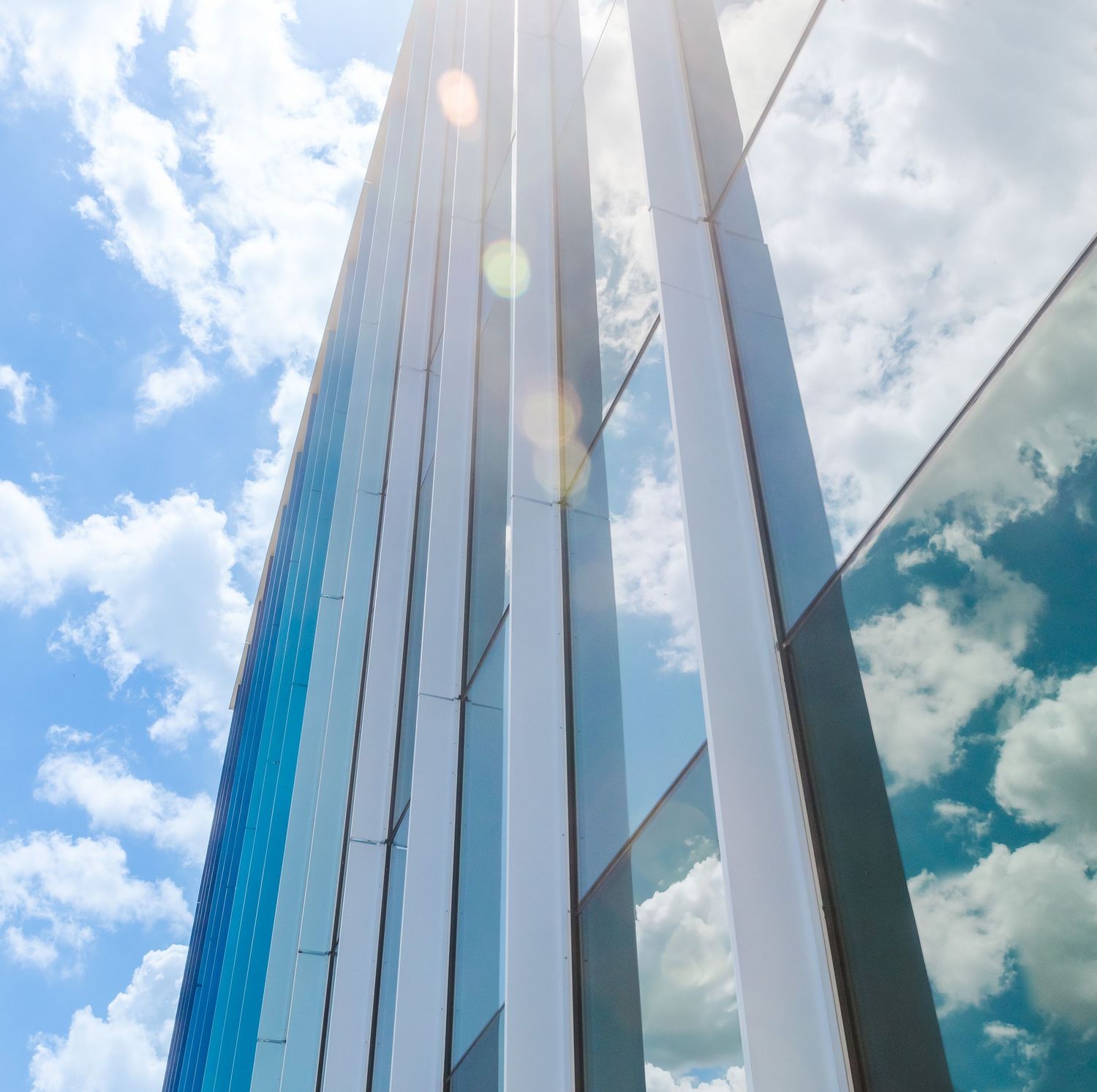Modern glass skyscraper facade with tall vertical lines reflecting blue sky and white clouds on a sunny day.