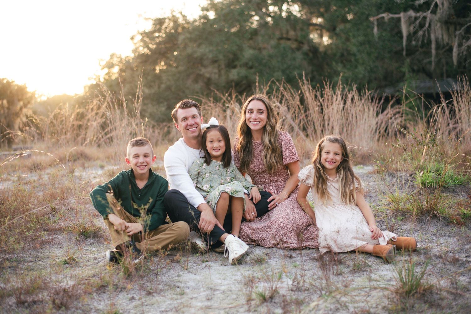 Family poses together on sandy beach during golden hour sunset photoshoot while wearing coordinated neutral outfits.