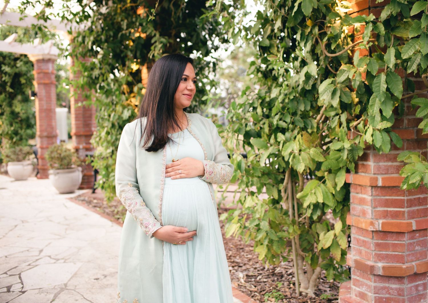 Long-haired figure wearing a light blue dress stands gracefully in a garden setting with brick pillars and climbing vines.