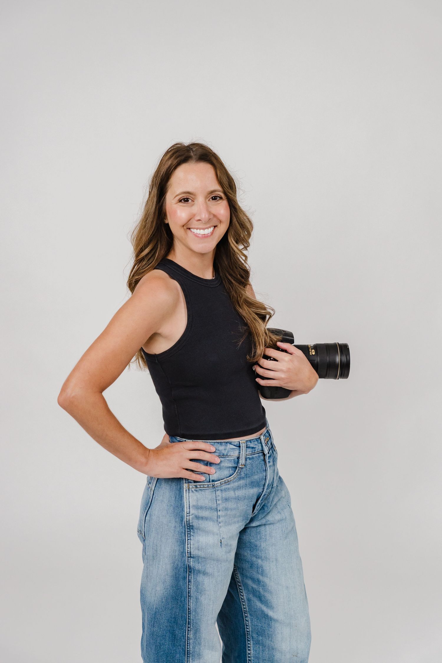 Professional photographer wearing black tank top and jeans holds camera while posing against light gray backdrop.