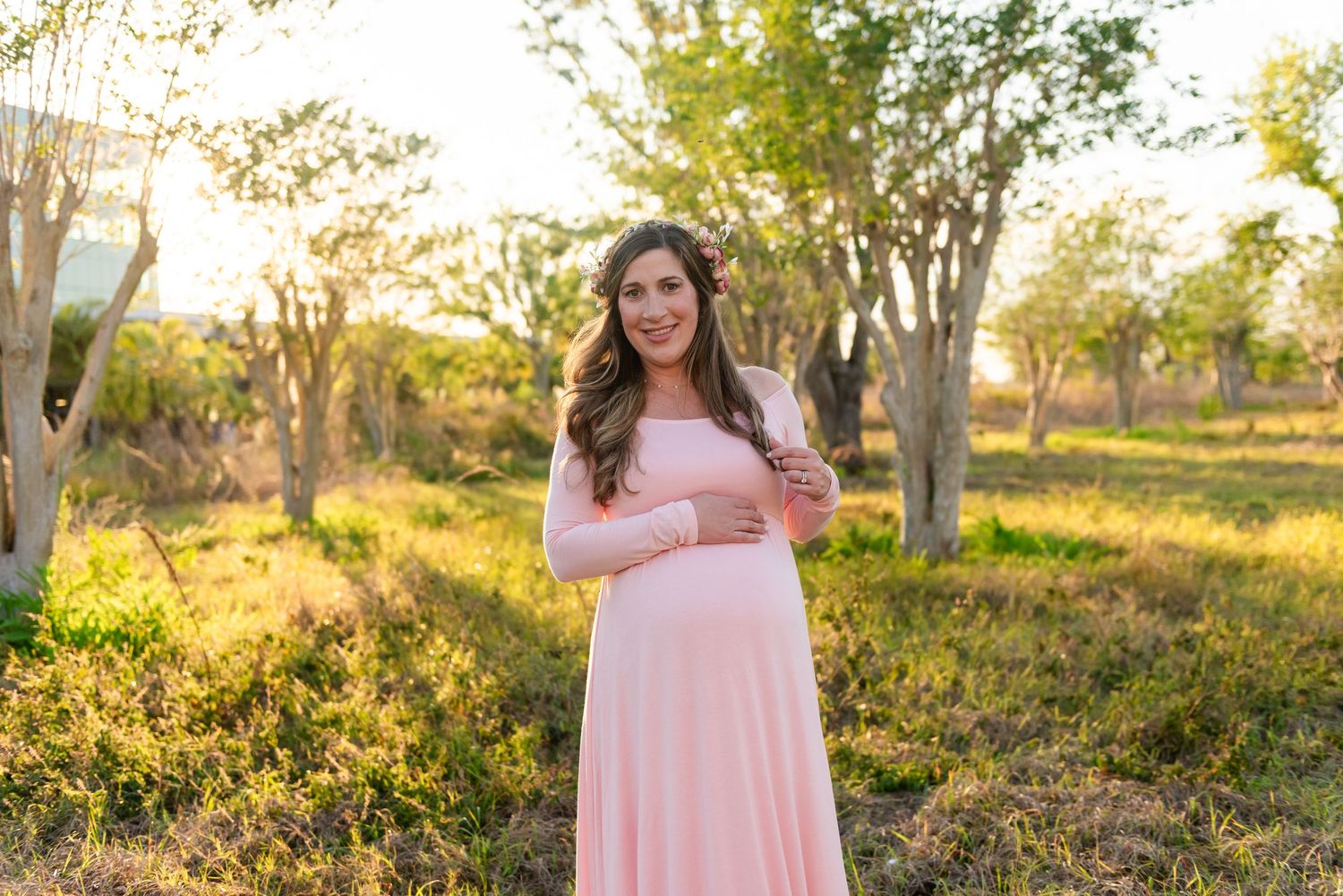Person in a flowing pink dress poses outdoors at golden hour in a natural setting with sunlit trees in the background.