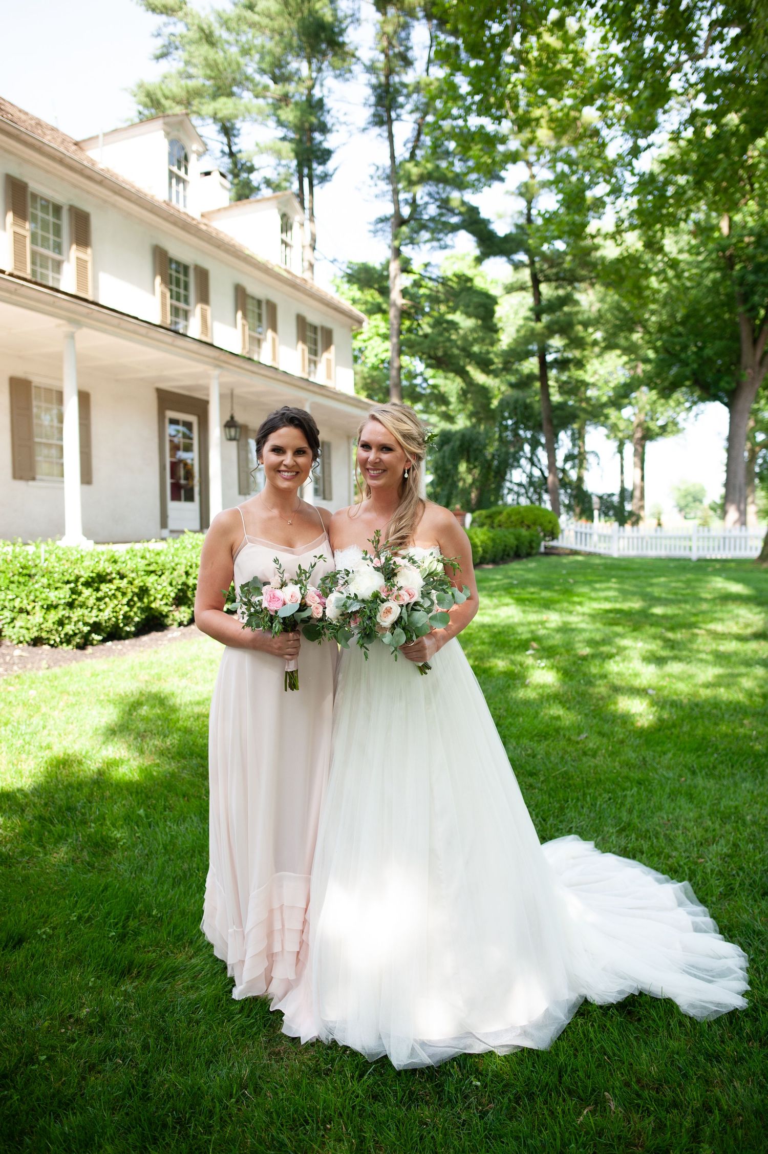 Two figures in blush and white formal gowns pose with bouquets in front of a stately white mansion.