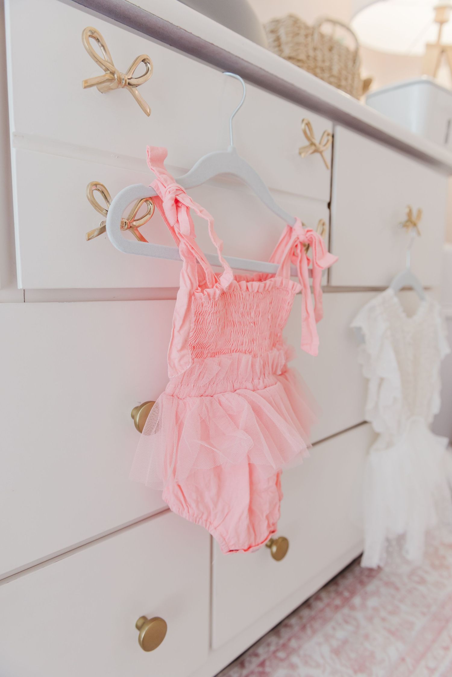 Pink ruffled baby romper hanging on white dresser with gold bow drawer pulls.