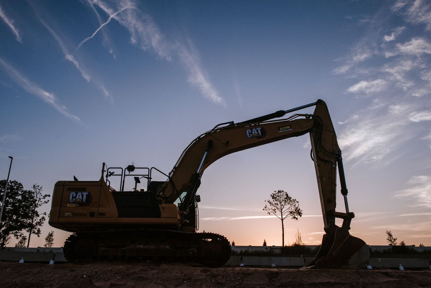 Silhouette of a CAT excavator against a dramatic sunset sky with wispy clouds and distant trees.
