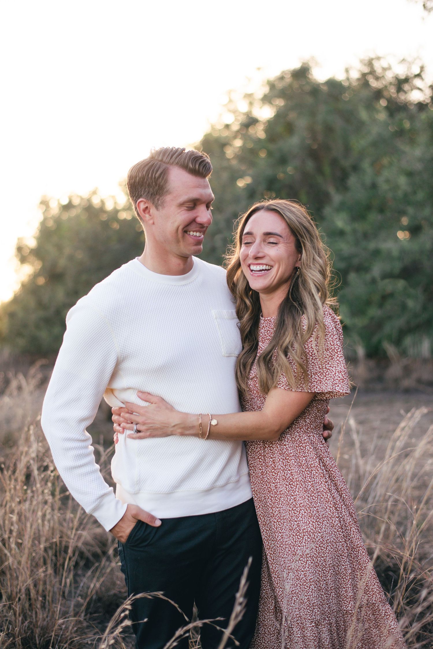 A happy couple laughing together in a golden field during sunset wearing white and pink outfits.