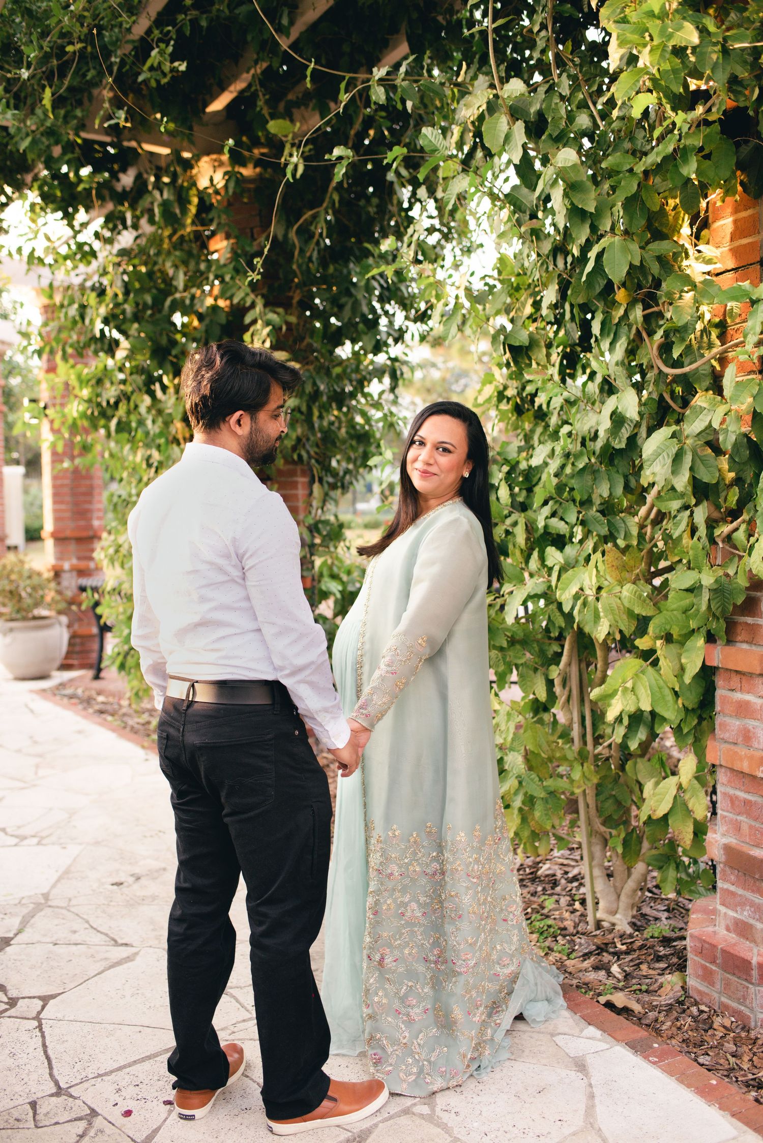 Two people holding hands under a leafy green arbor on a brick paved pathway during an outdoor engagement shoot.