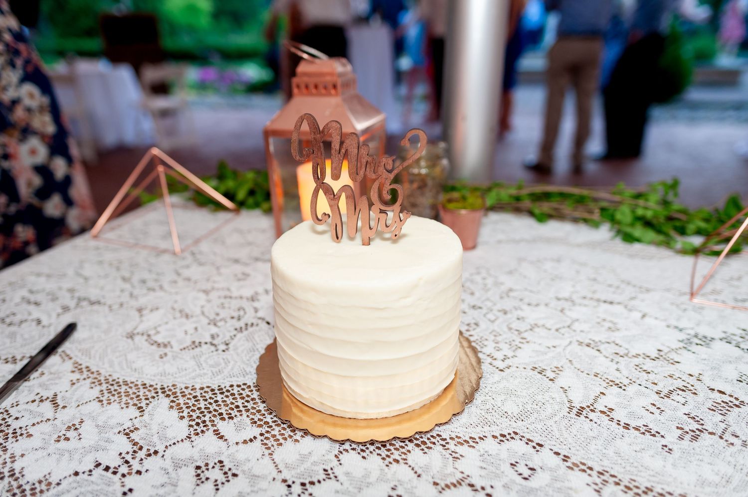 Small white wedding cake with wooden cake topper displayed on lace tablecloth at outdoor reception.