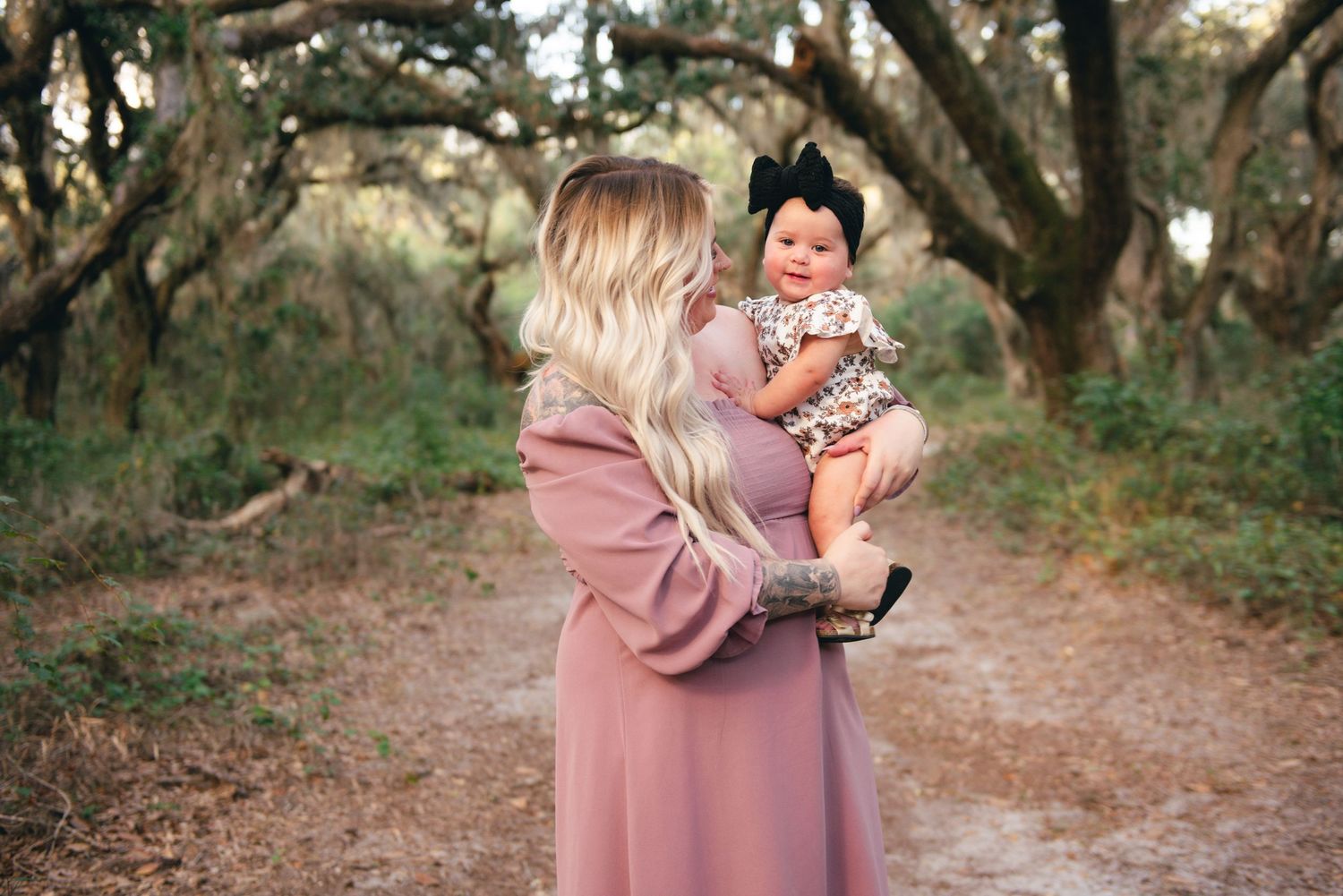Parent in mauve dress holding baby on wooded trail during photo session.