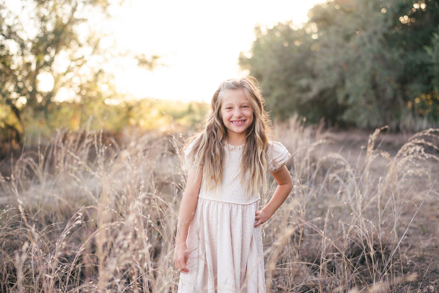 A person in soft pink clothing stands in golden tall grass during sunset.