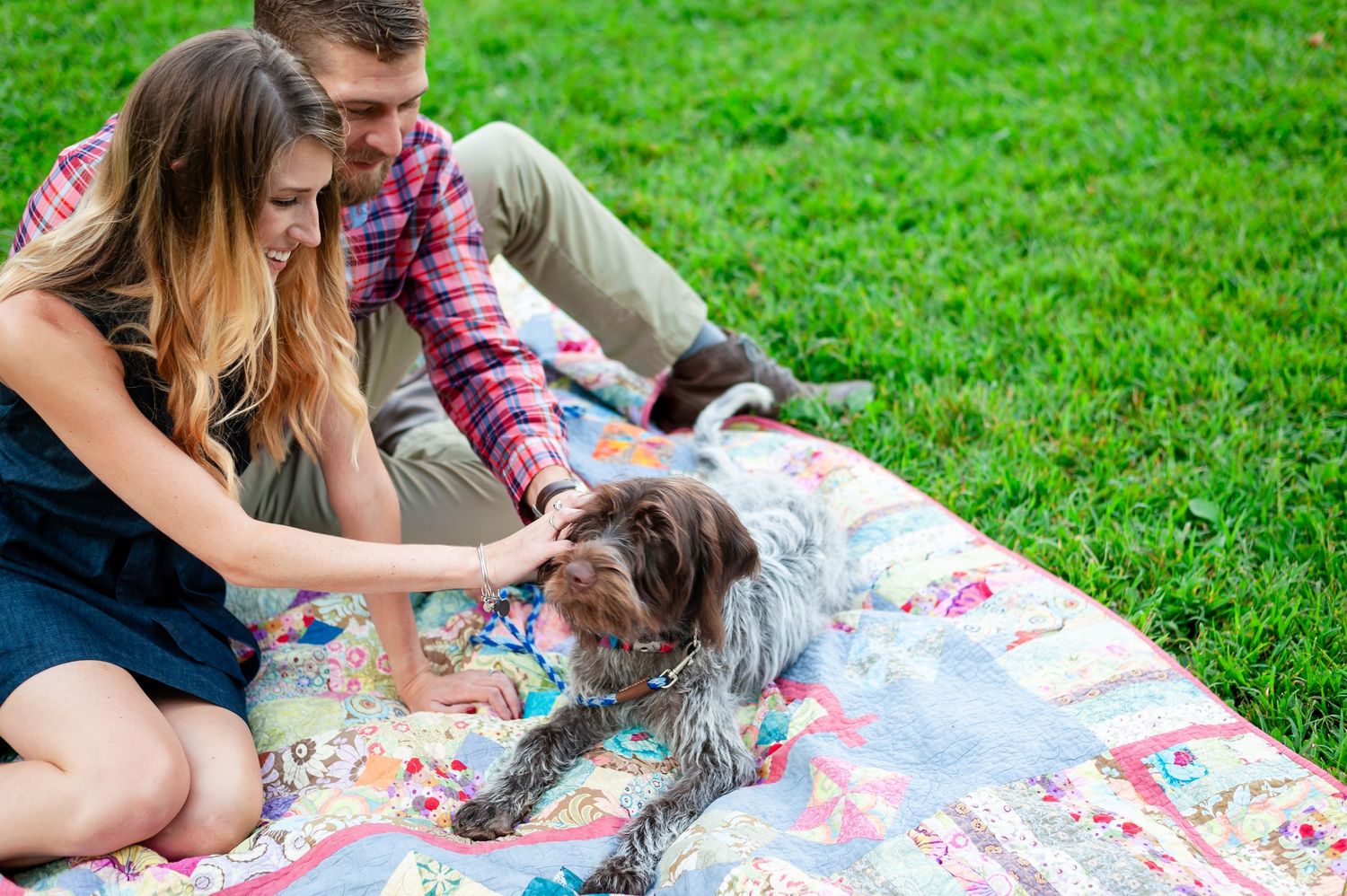 Couple plays with their dog while relaxing on floral blanket in green park.