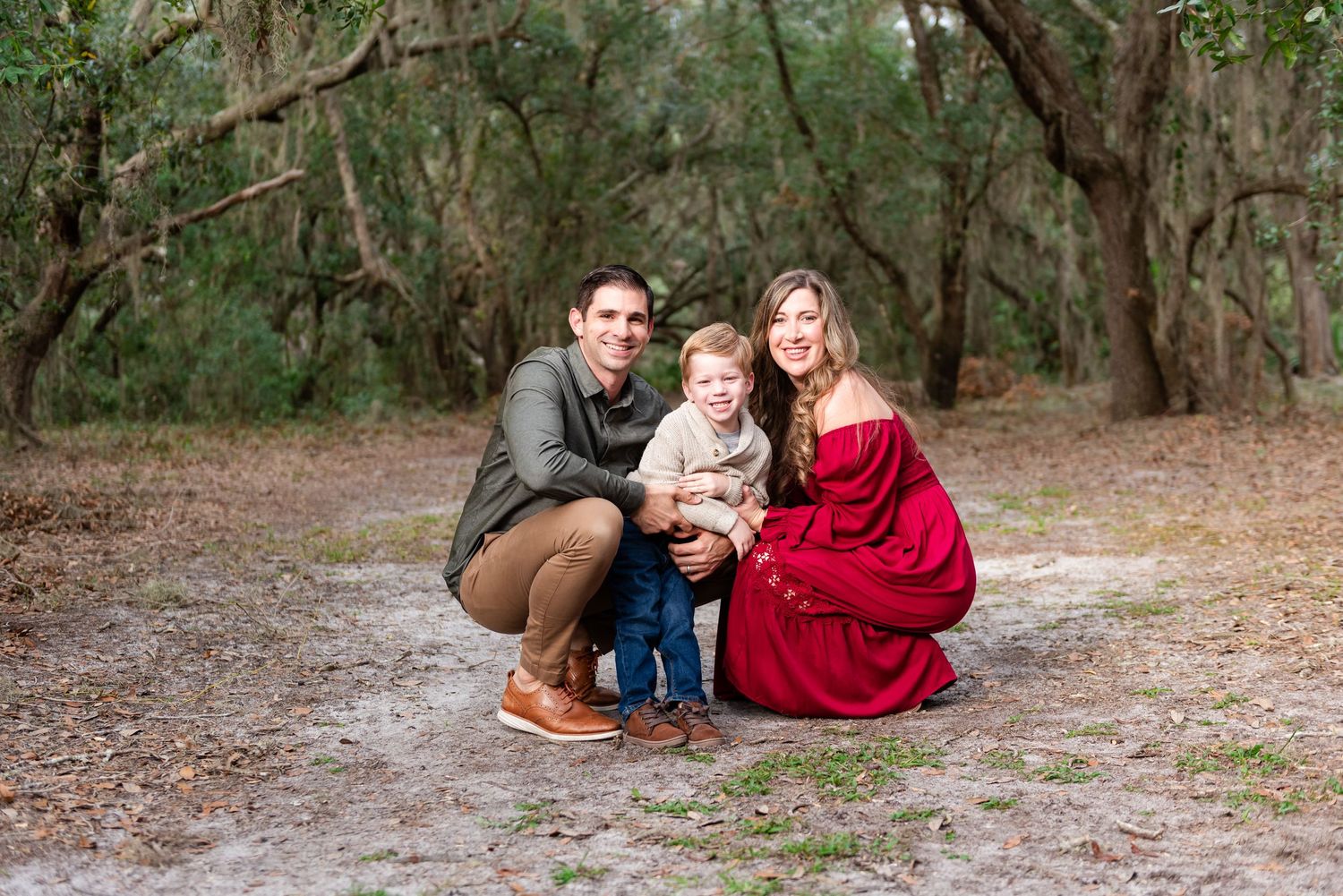 Two people in red and navy outfits sit together on a sandy path in a wooded outdoor setting.
