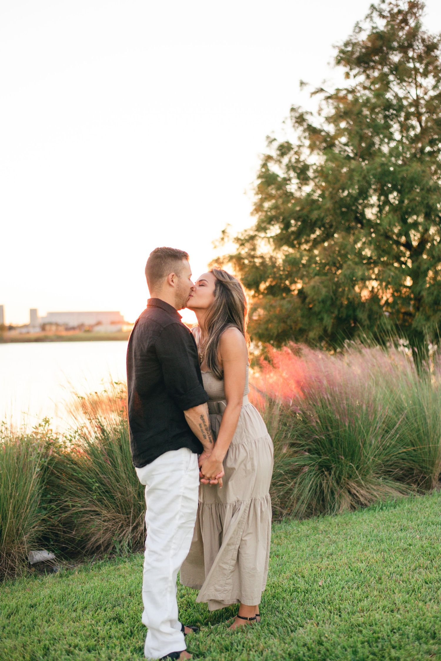 A couple kisses while standing on grass near a lake during a beautiful sunset.
