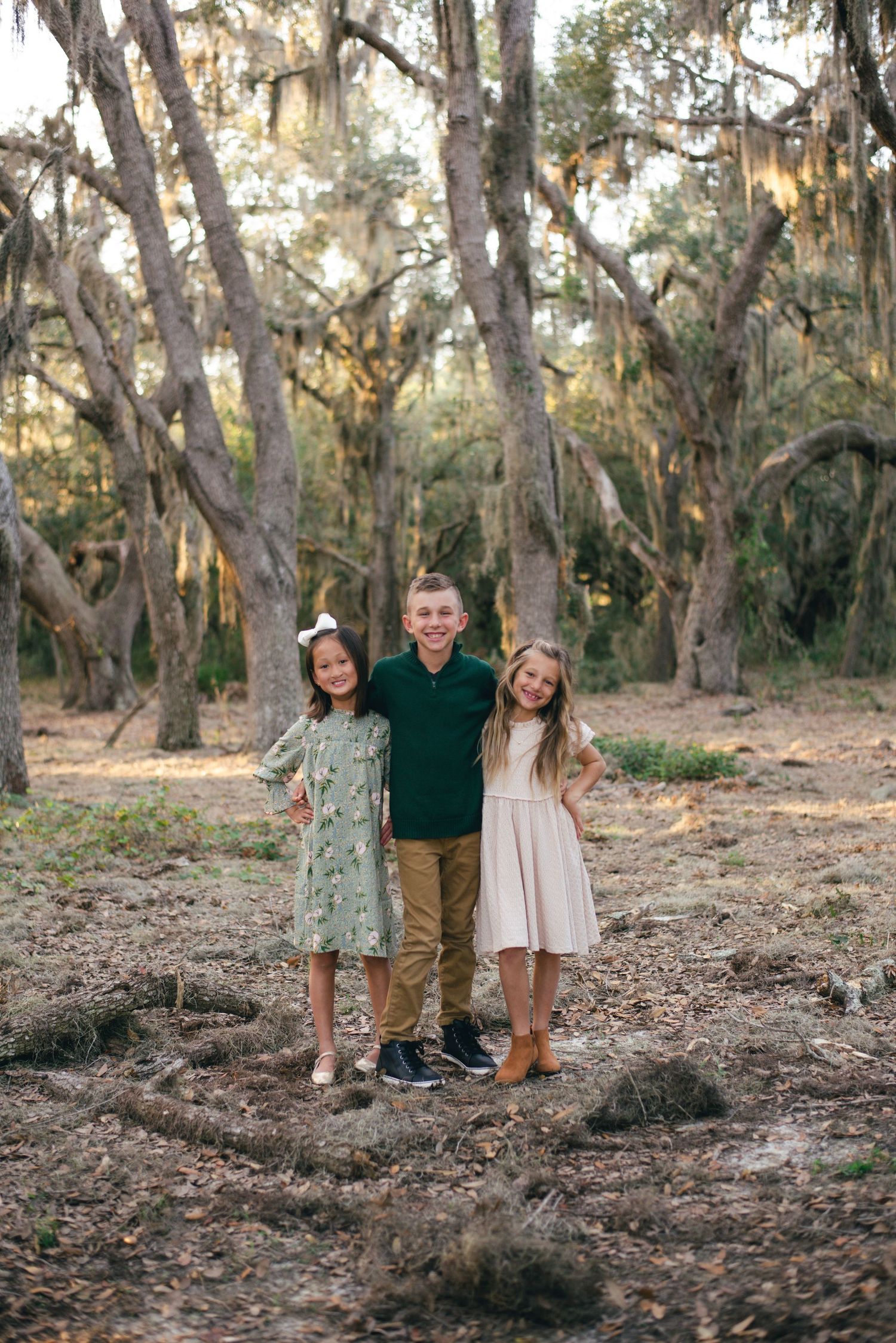 Three young children stand together on leaf-covered ground with Spanish moss draped trees in the background.