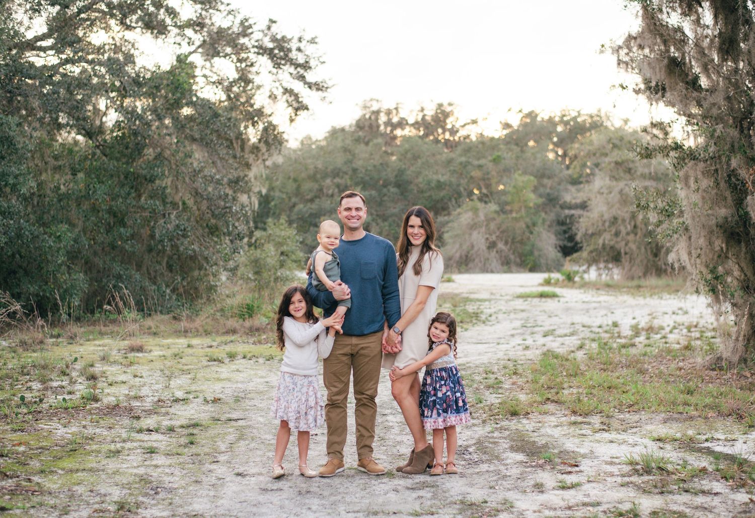 A family group stands together on a dirt path surrounded by trees and Spanish moss in a natural outdoor setting.