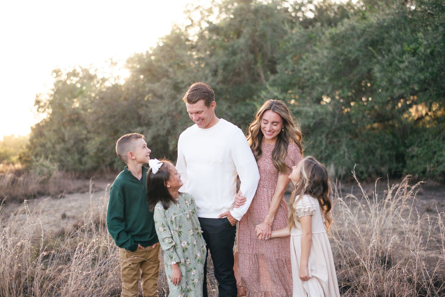 A family in casual attire stands together in a golden field at sunset with trees in the background.