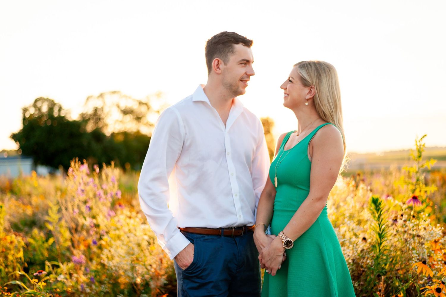 Couple in white shirt and green dress stand together in wildflower field at sunset.