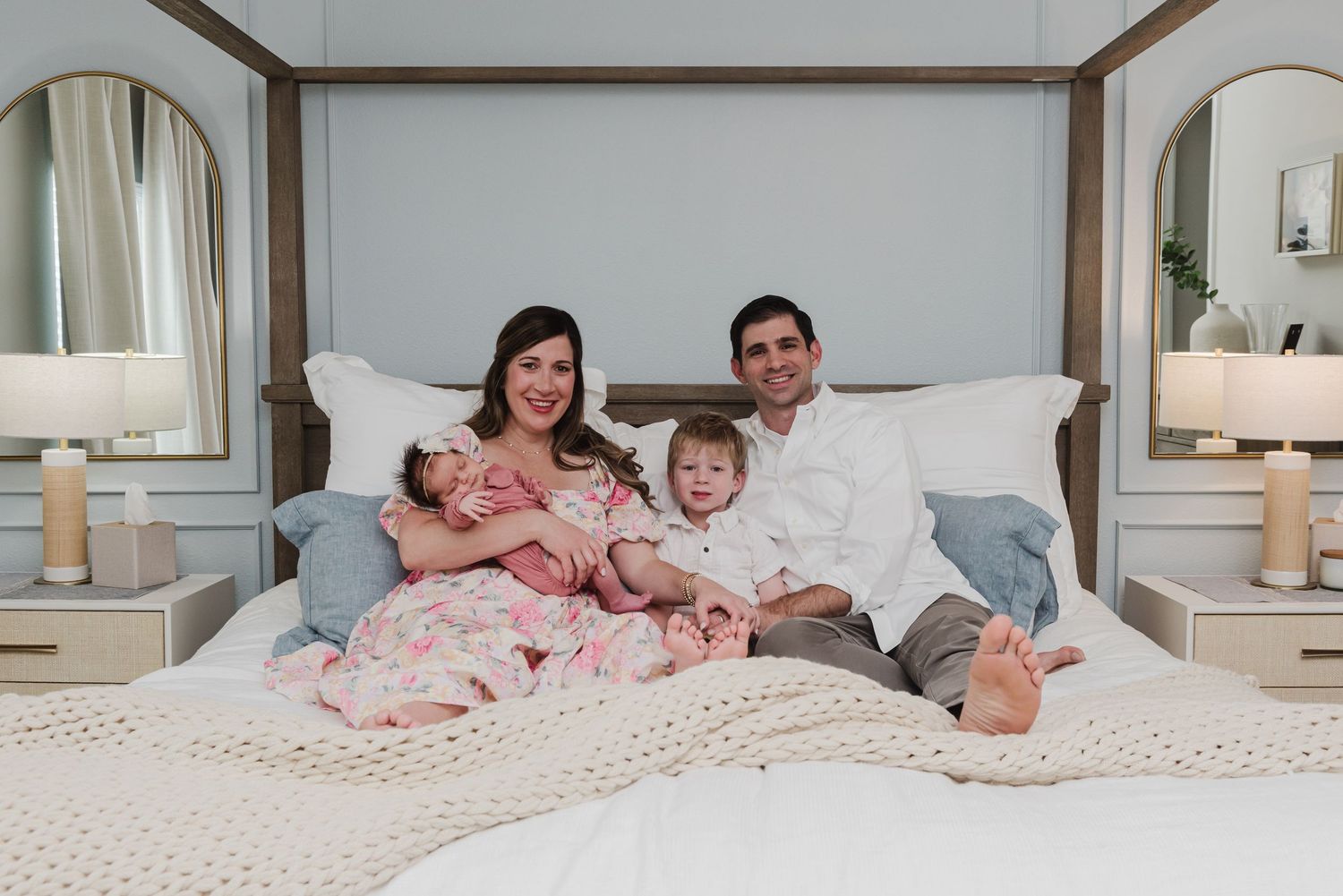 Family of four relaxes together on white bed with matching decor and bedside lamps.