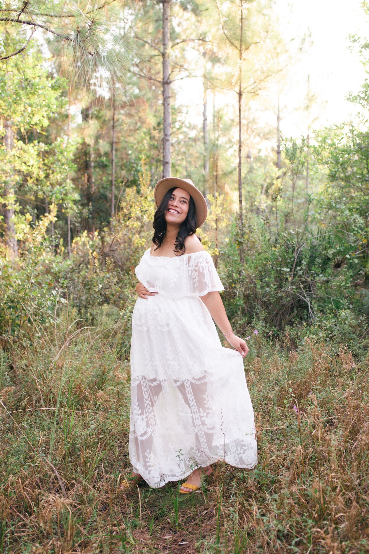 A white lace off-shoulder dress with sun hat in a dreamy forest setting with golden sunlight filtering through trees.