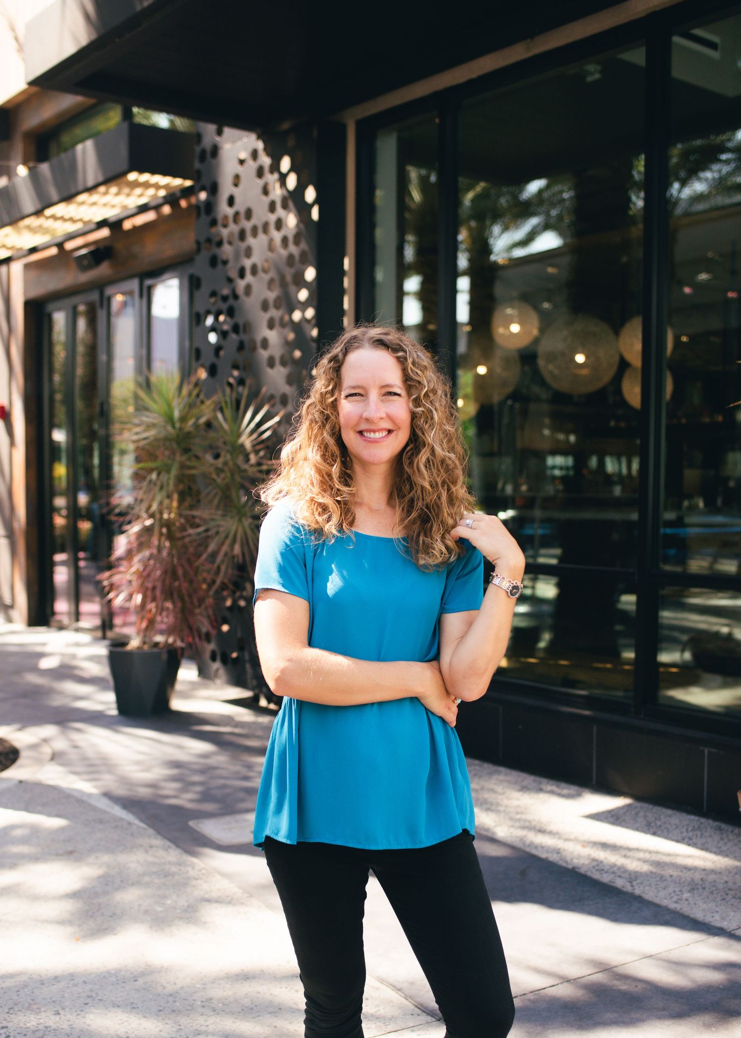 Woman in flowing blue top standing outside modern retail space with palm trees.