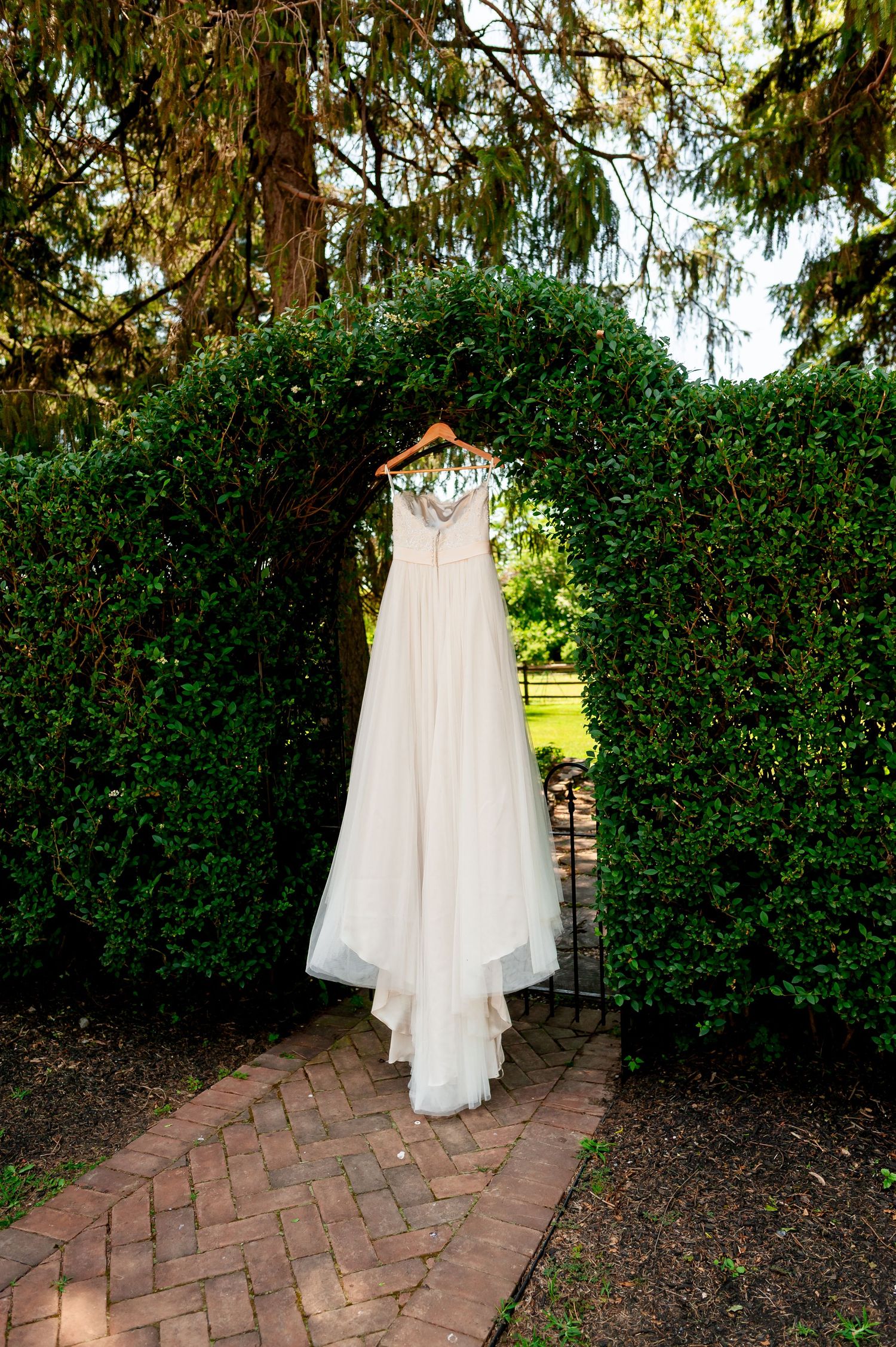 White wedding dress hanging on display against lush garden hedge and brick pathway.