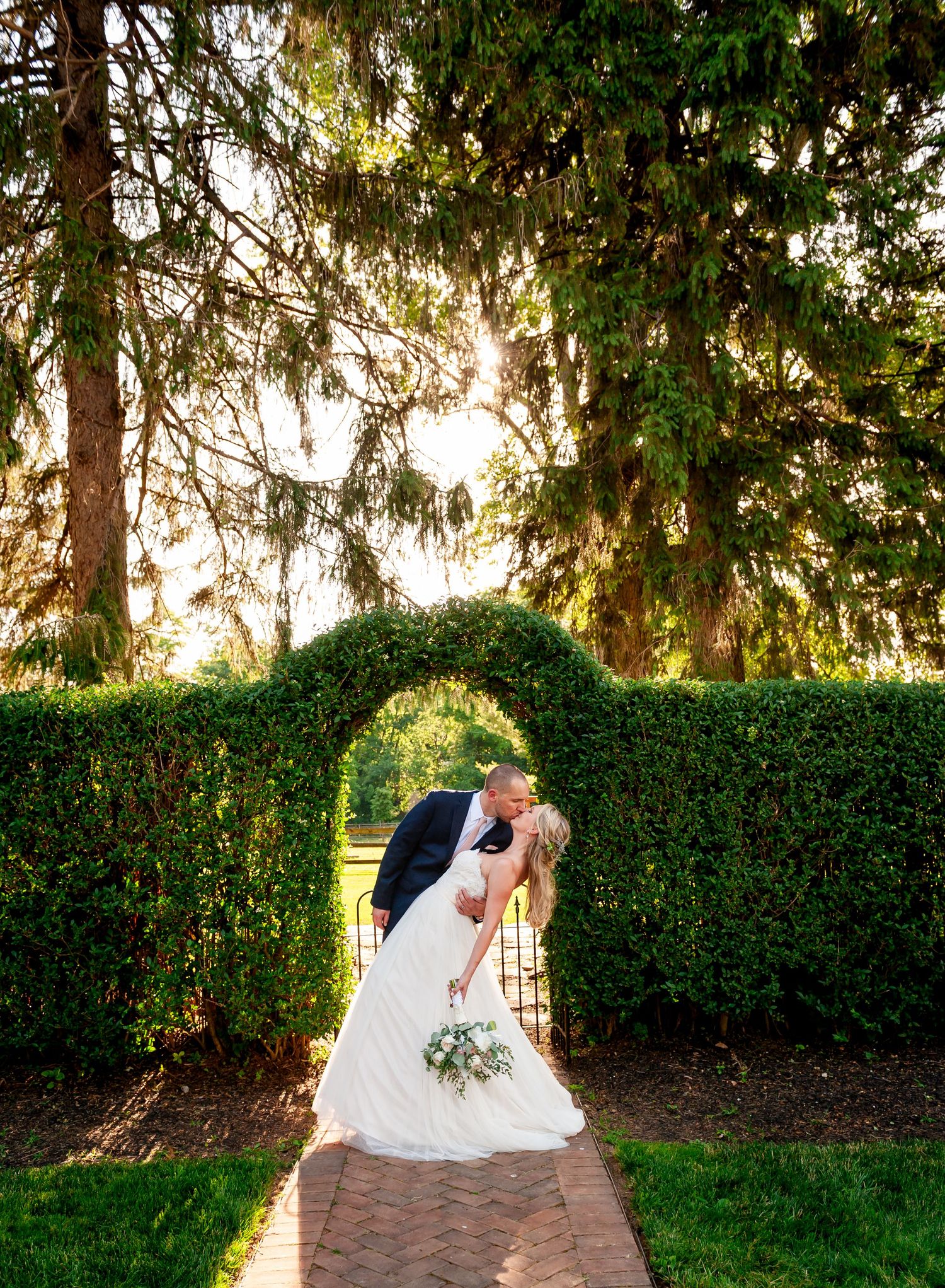 Romantic wedding portrait under hedge archway with sunlight filtering through trees.