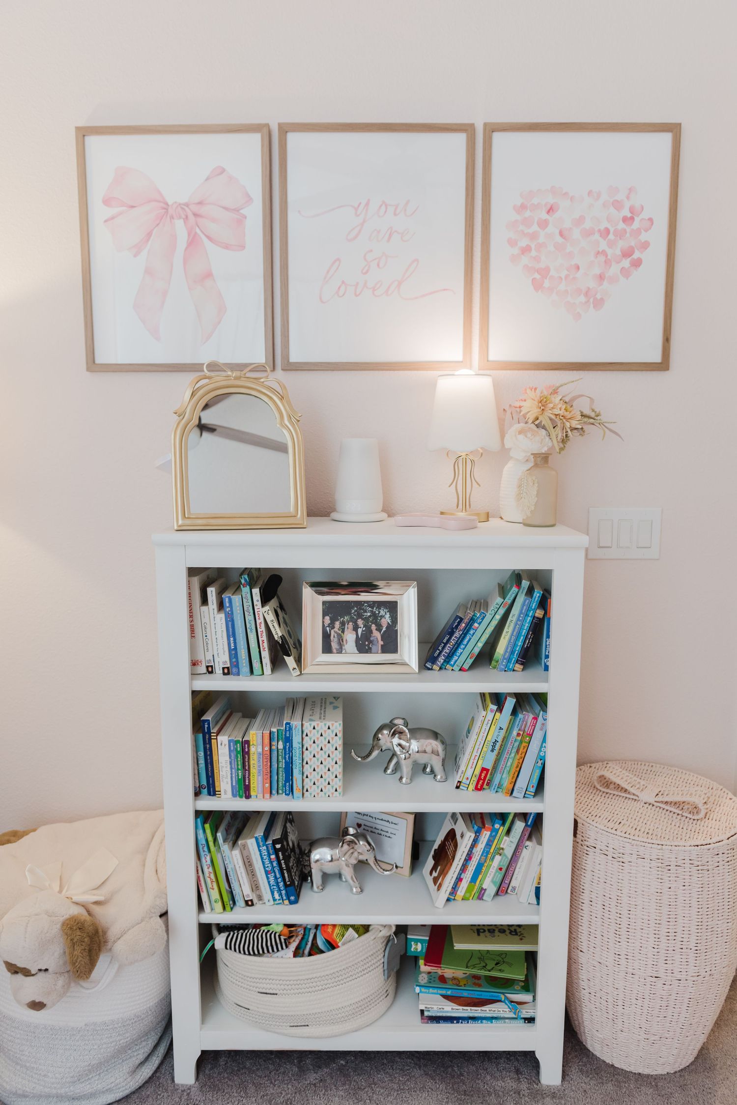 White bookshelf with pink wall art and decorative items in a nursery setting.