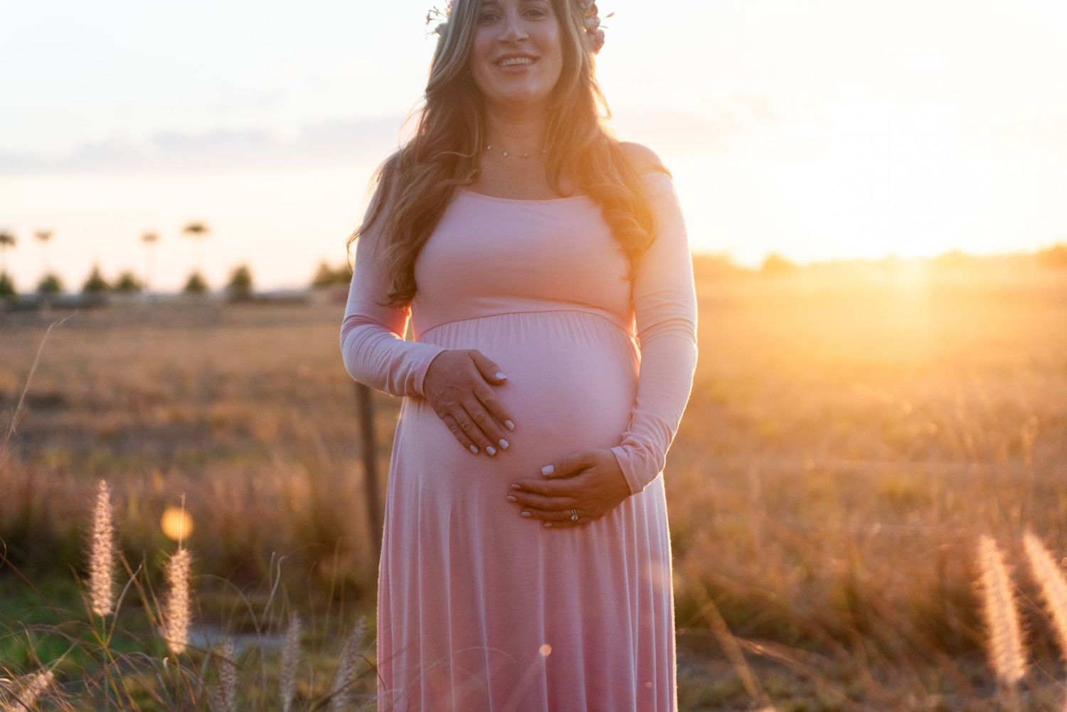 A glowing maternity portrait shows a person in pink cradling their belly among tall grasses at sunset.