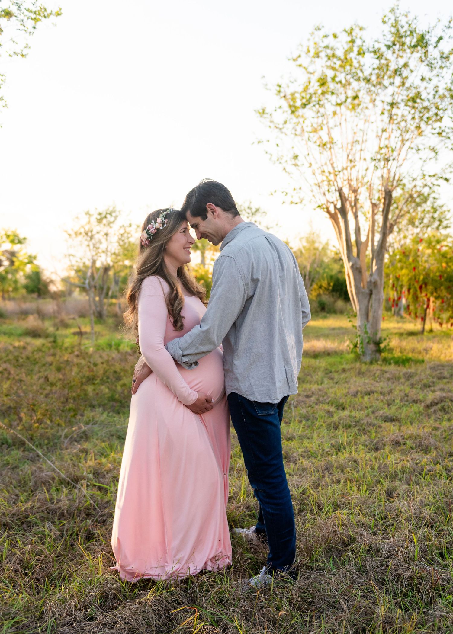 A couple stands together in a field during sunset, with the person wearing a flowing pink dress.