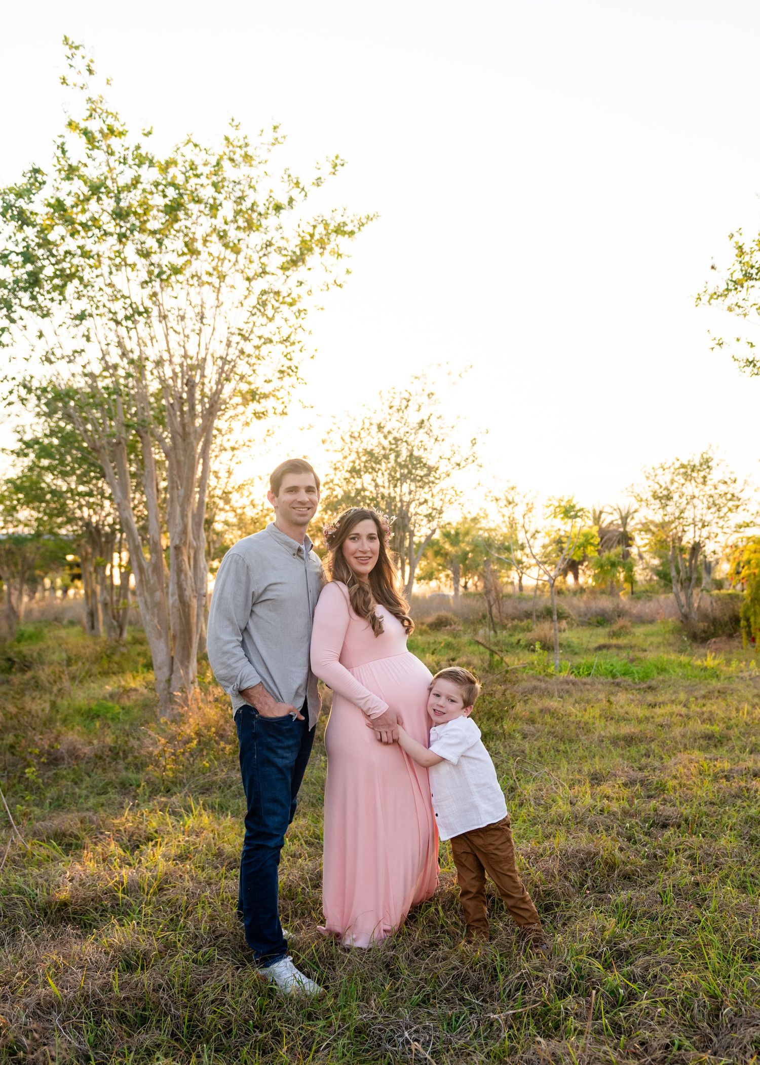 A family stands together in an outdoor field during golden hour sunset with trees in the background.