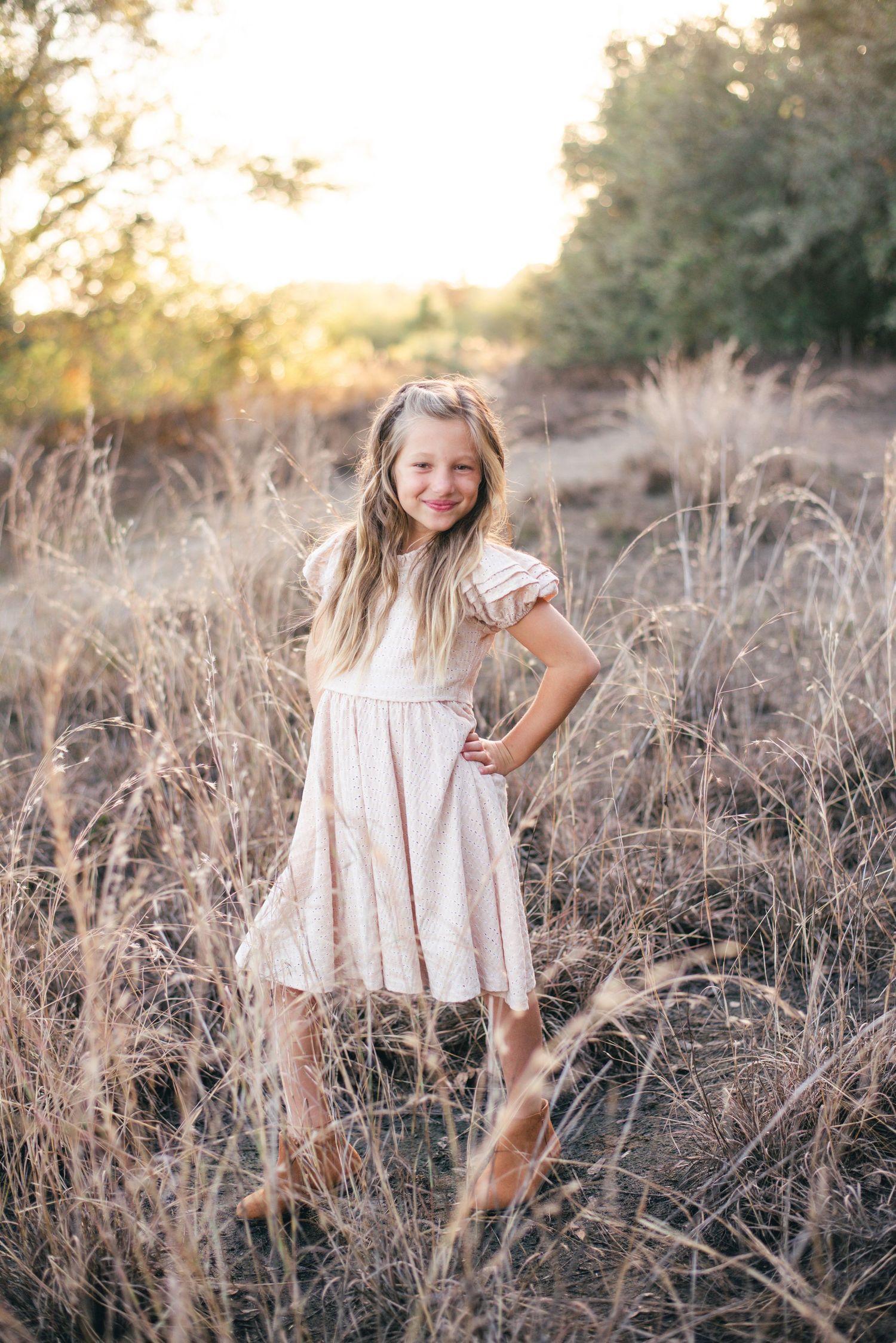 Young child in cream-colored dress poses playfully in tall grass at sunset with golden backlight.