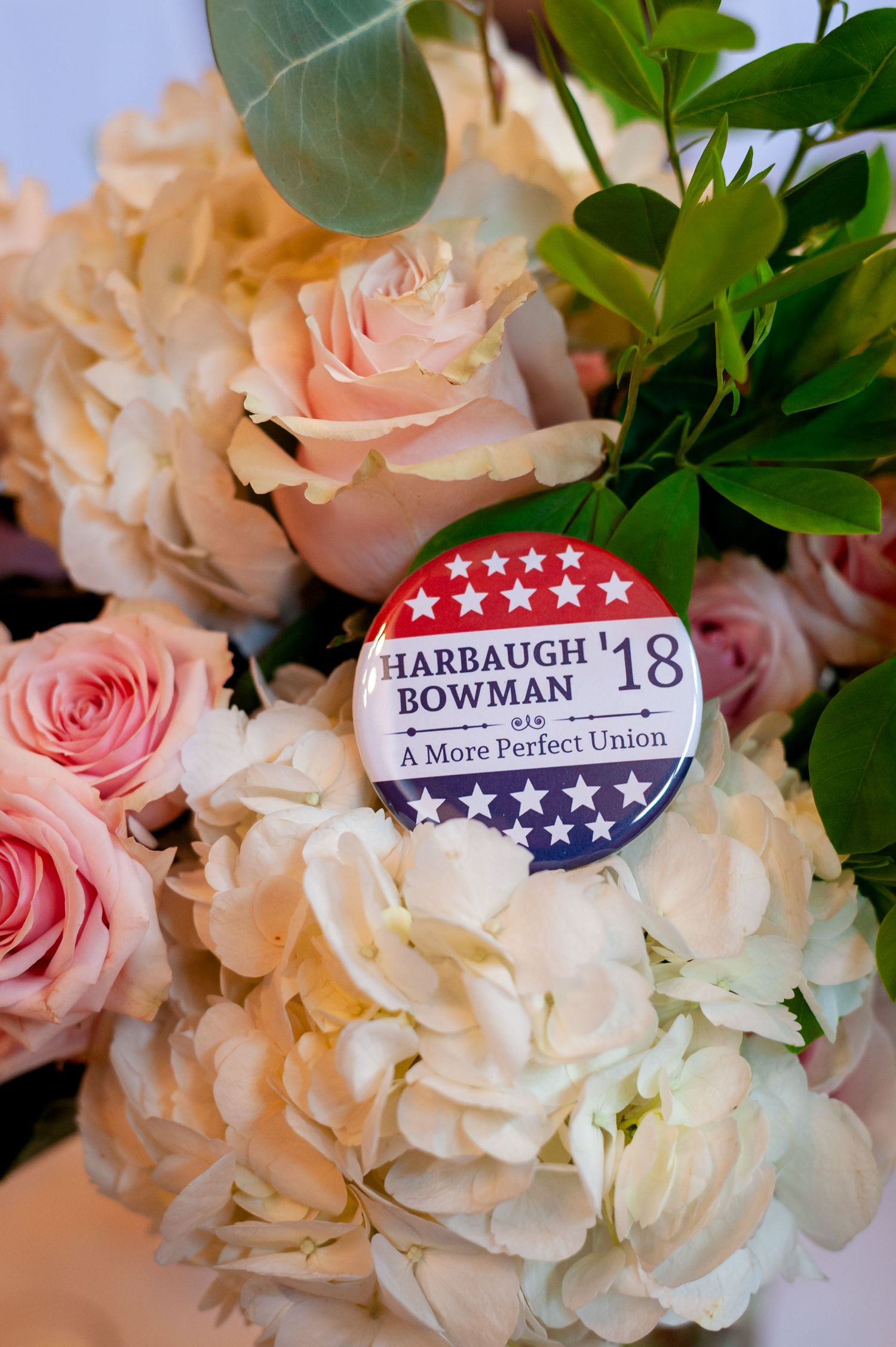 A political campaign button among pink roses and white hydrangeas in a floral arrangement.