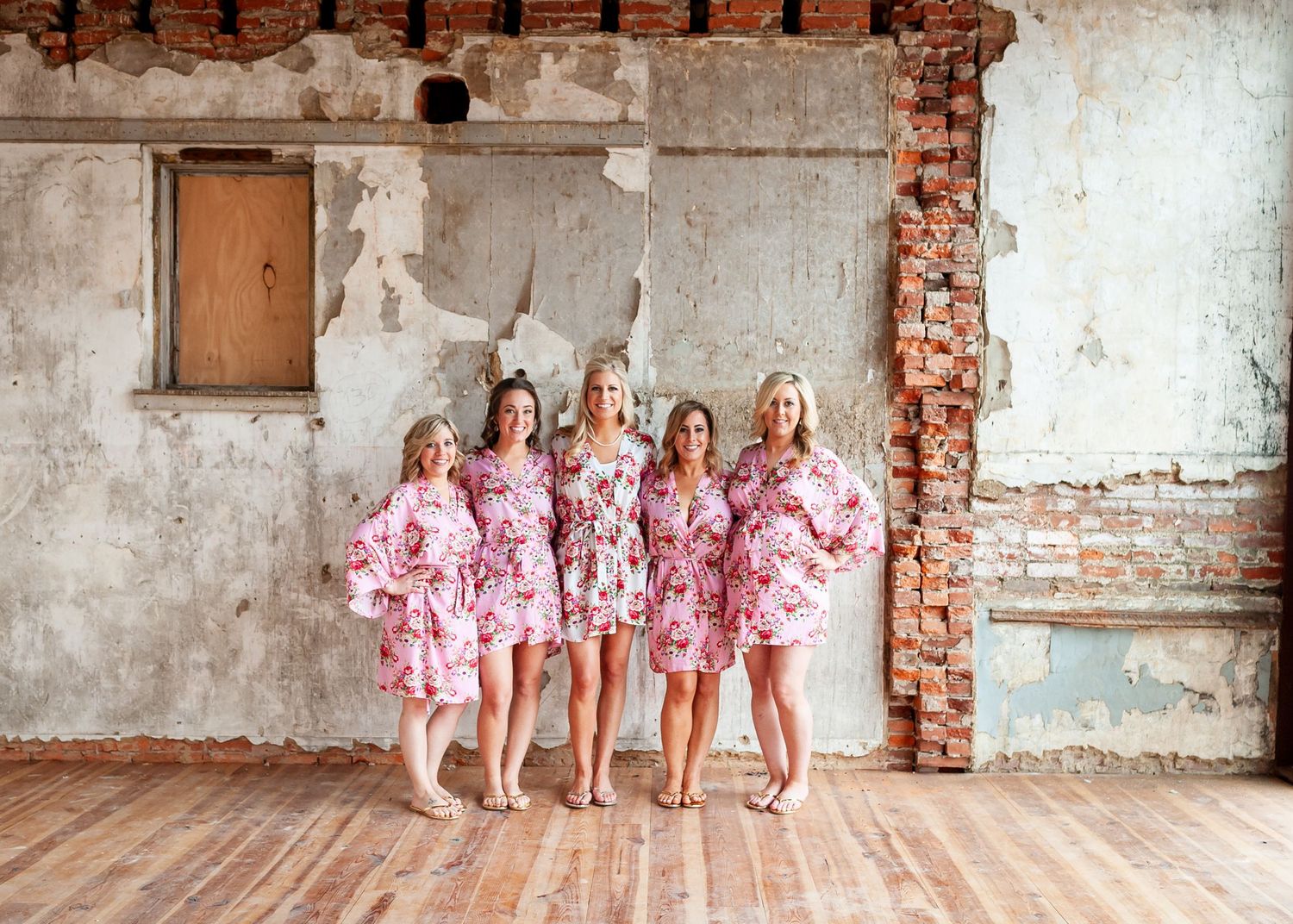 Group of bridesmaids in pink floral robes posing against industrial brick wall.