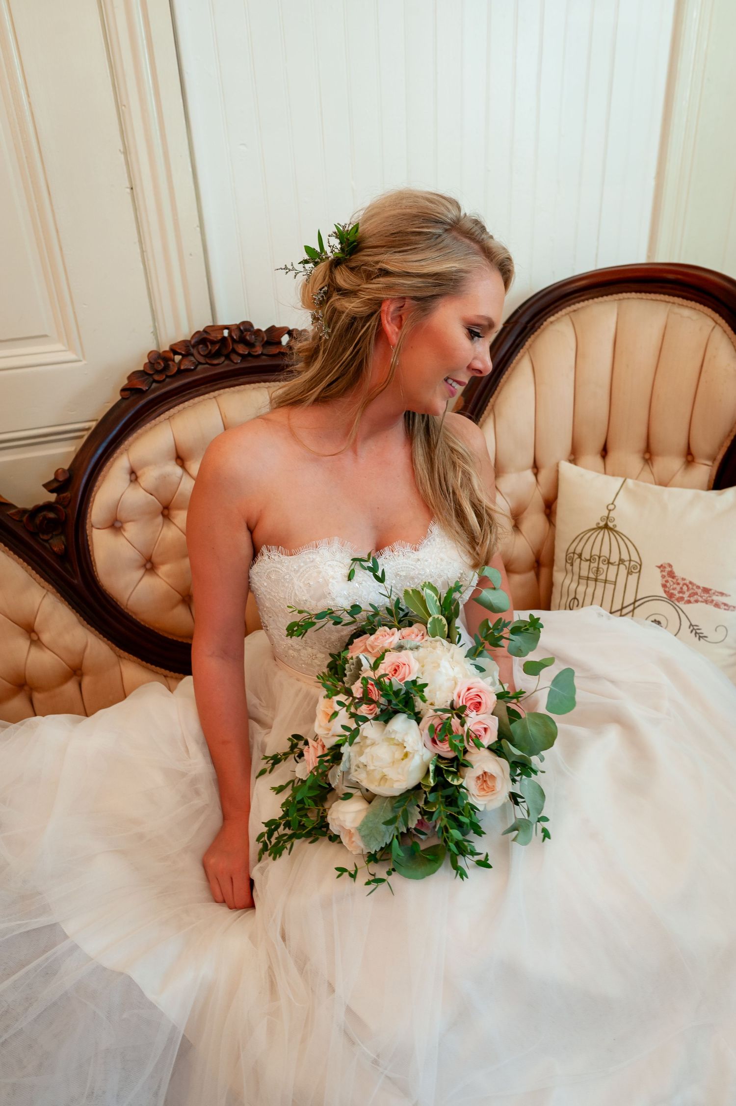 Bride in white strapless gown holding pink and white bouquet while seated on vintage sofa.