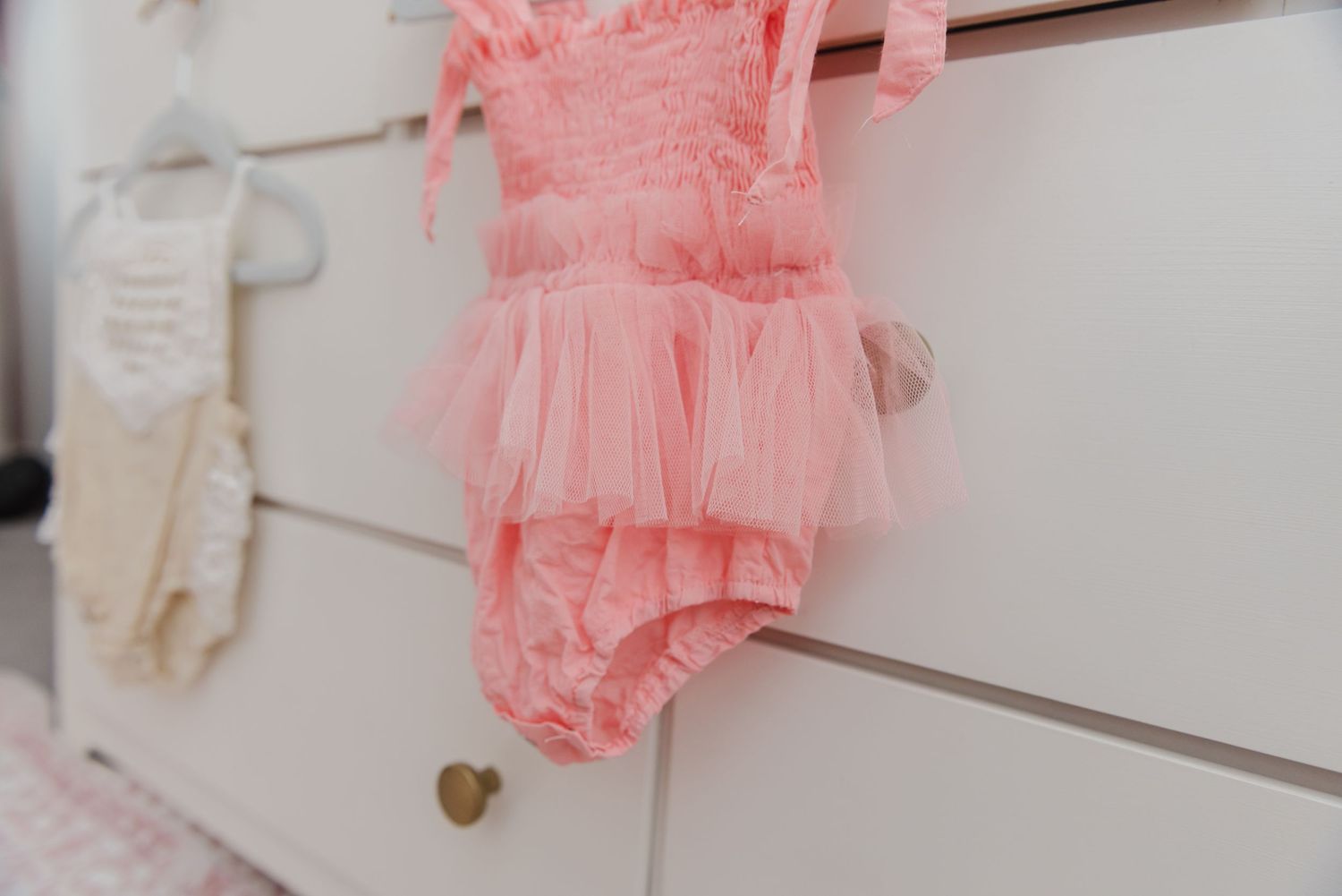 Pink ruched romper with tulle details hanging on white dresser drawer.