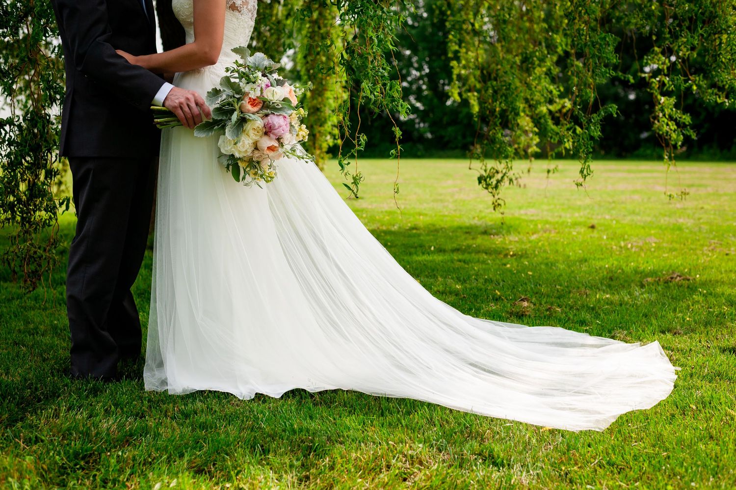 A flowing white wedding dress train extends across green grass in an outdoor setting.