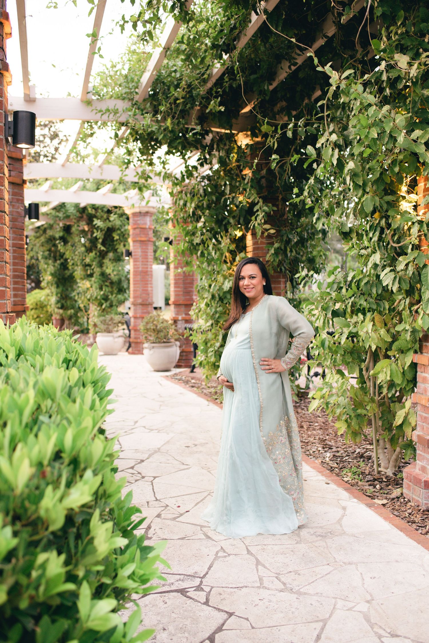 A person in a light blue maxi dress poses against a garden wall with lush greenery and a stone pathway.