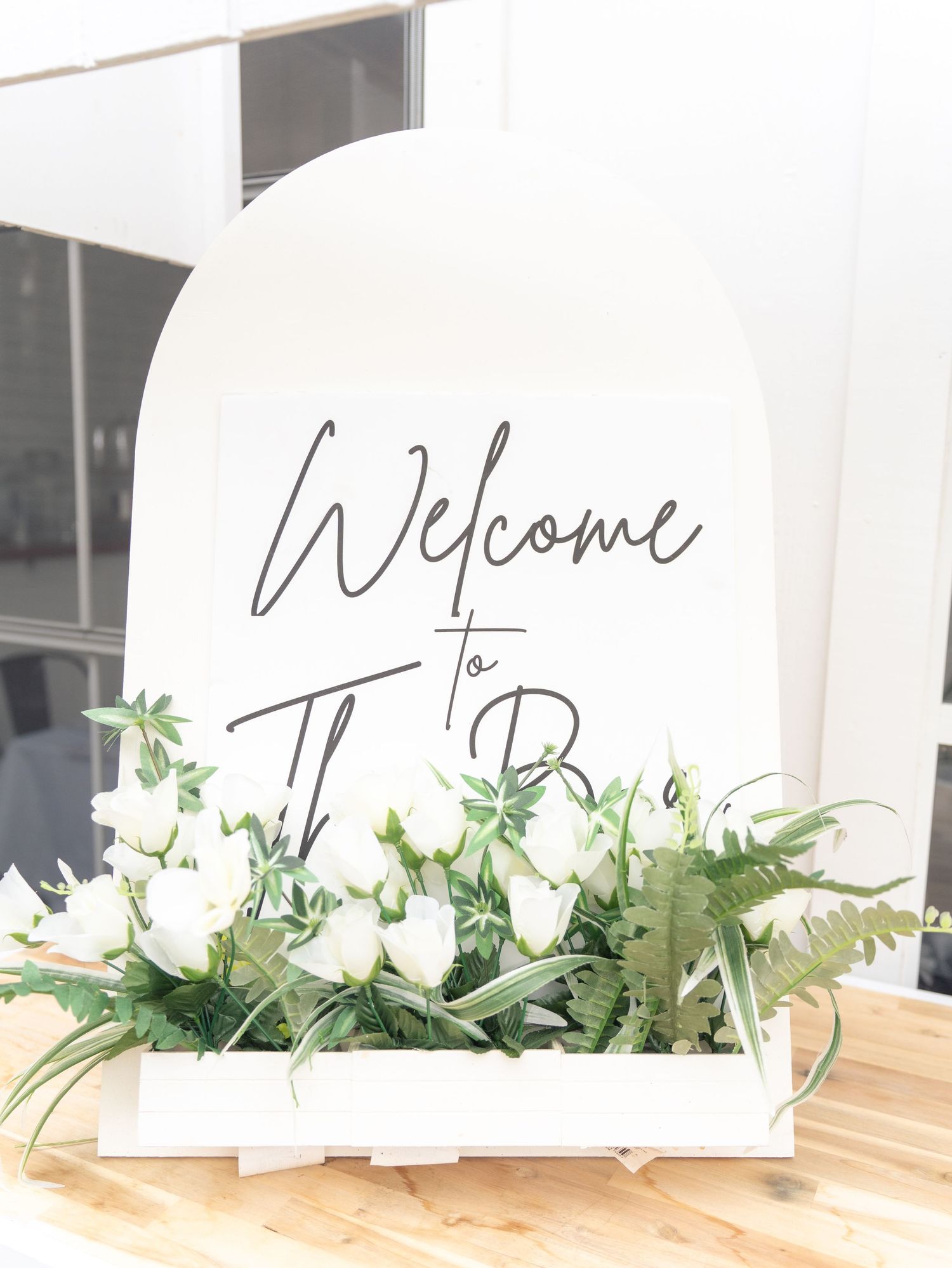 Welcome sign decorated with white roses and greenery on white curved backdrop.