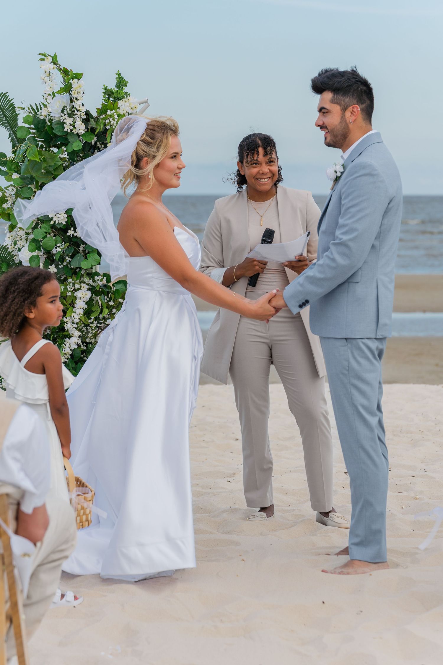 A beach wedding ceremony with white decor and greenery on a sunny day.