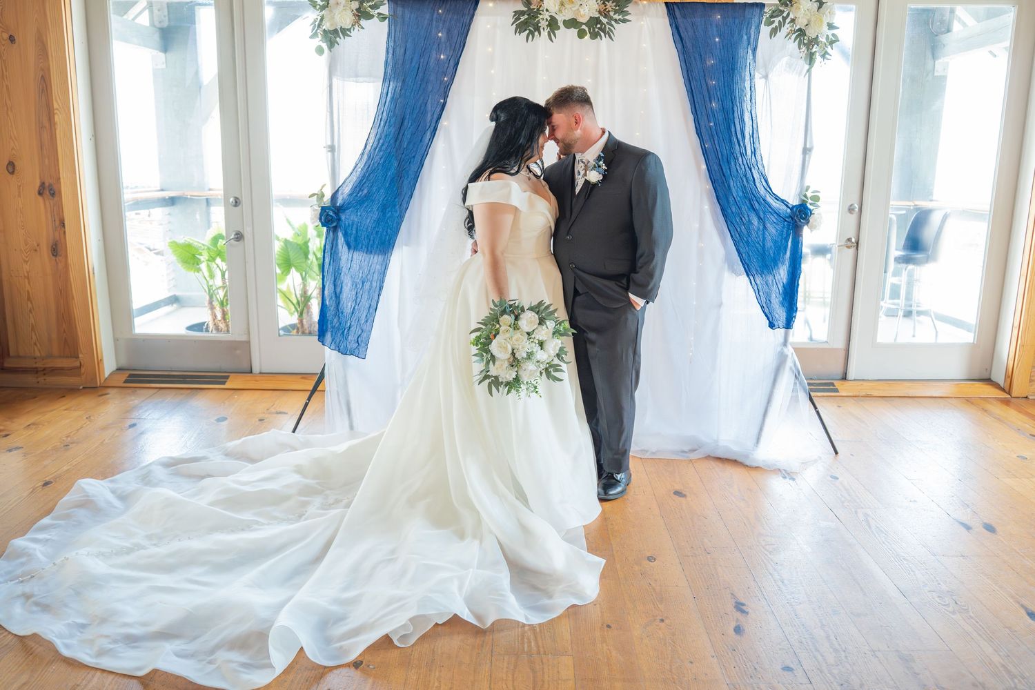 A wedding ceremony takes place in an elegant indoor venue with blue curtains and floral decorations.