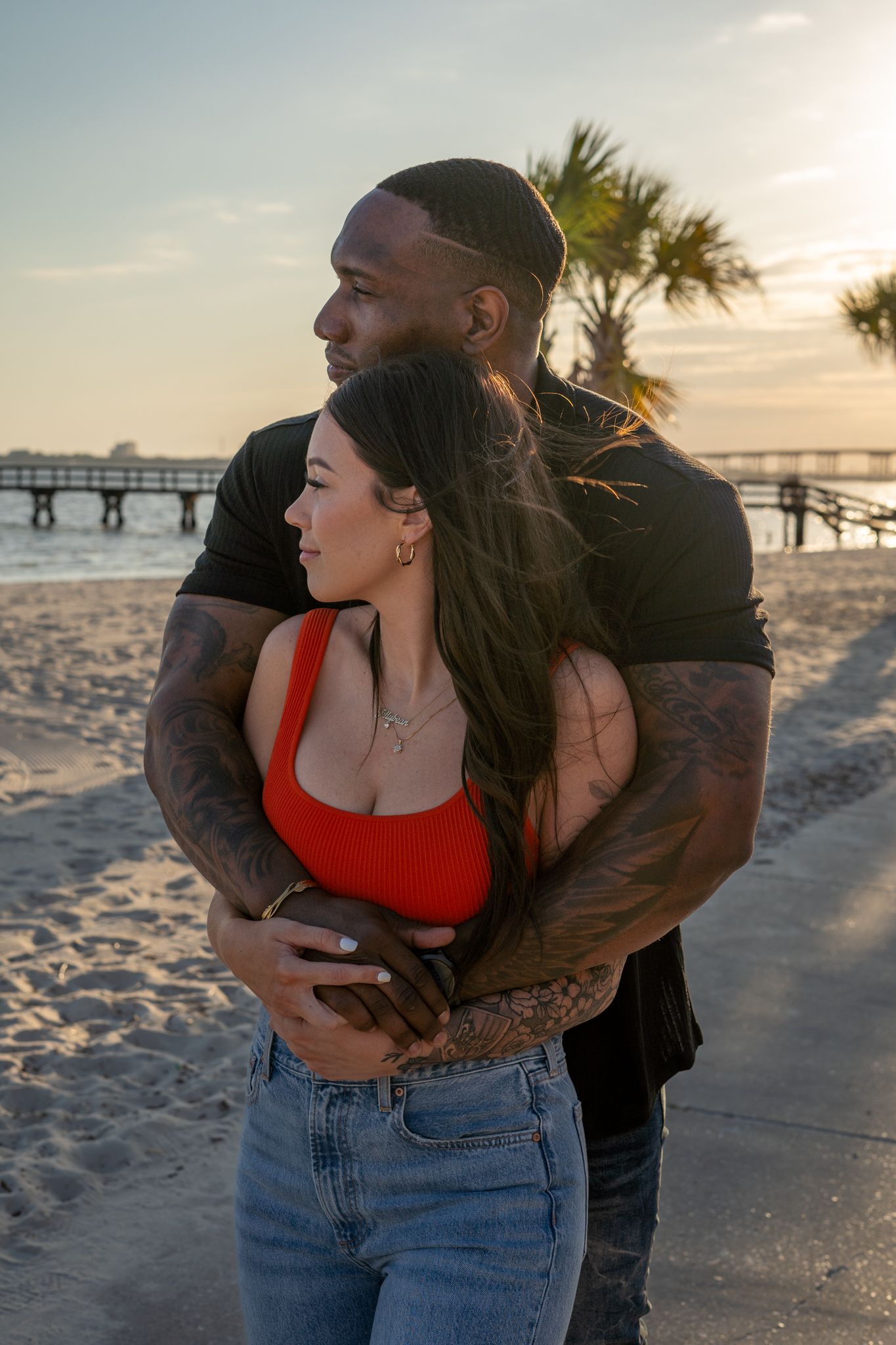 Couple embracing on a sandy beach with palm trees and pier in background.